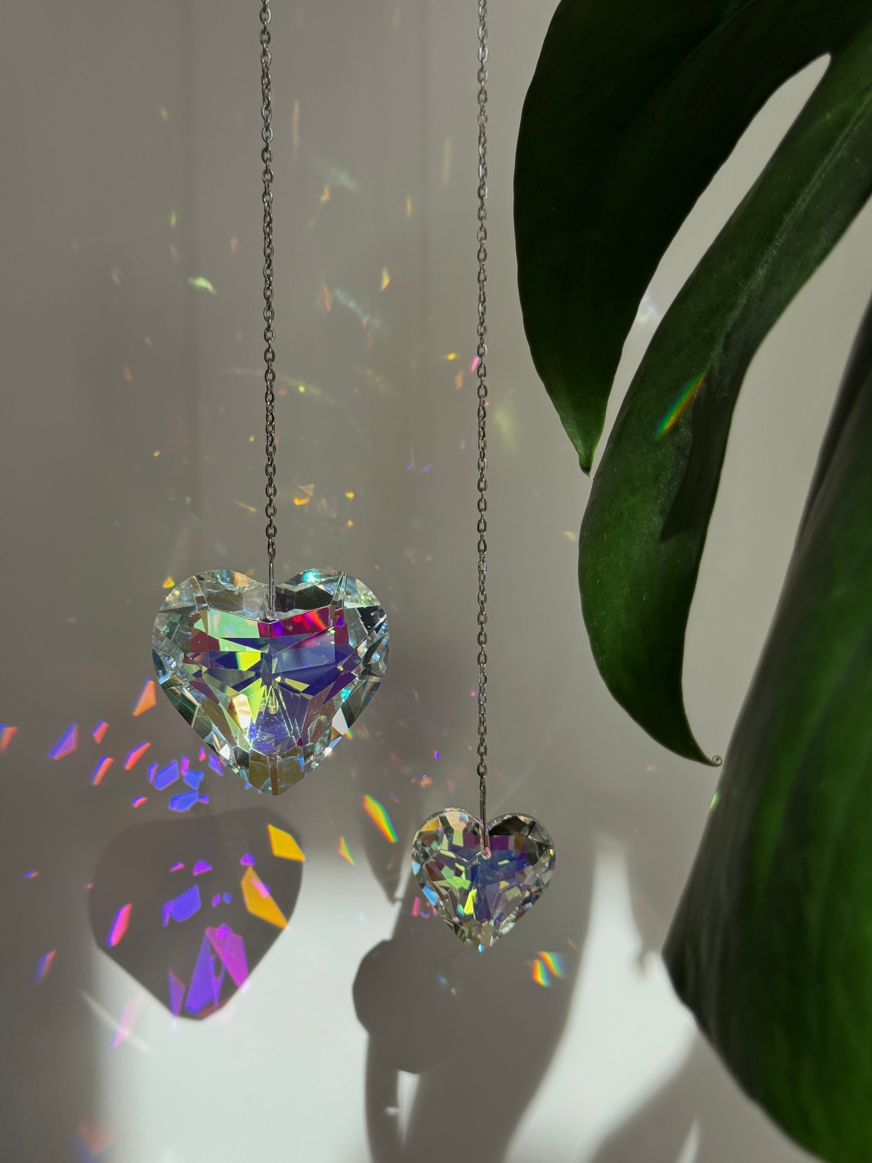Two iridescent heart crystals, 3cm and 5cm, reflecting sunlight into colorful spectral patterns. Part of the 'Sweet Reflection' collection, showing the stainless steel hanging hook and delicate chain.