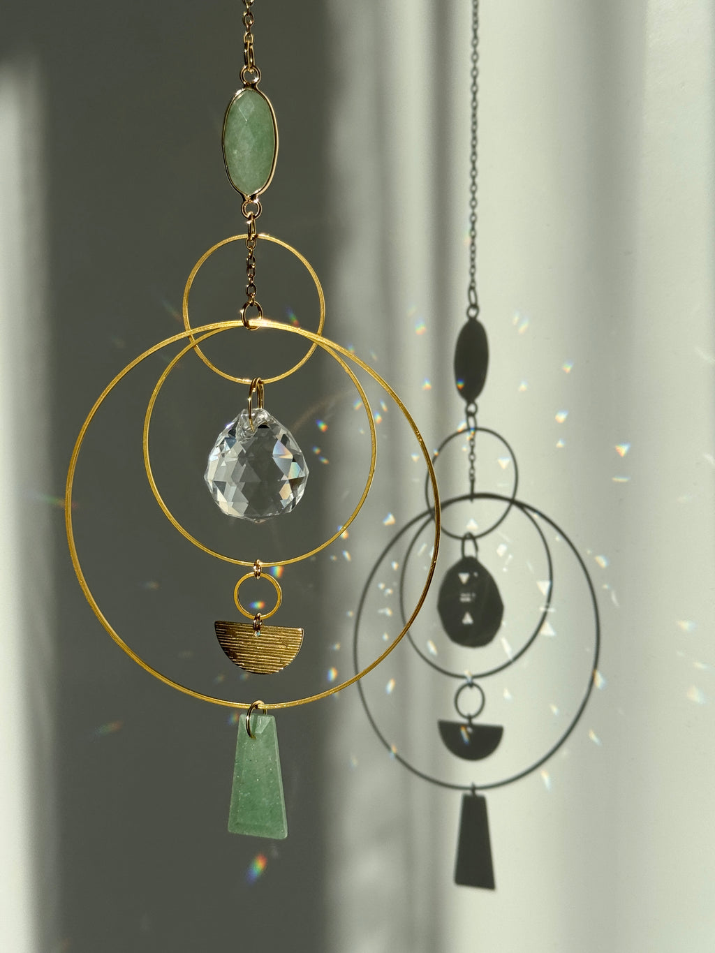 Vertical gold geometric suncatcher with three tiered hoops, a faceted crystal sphere, and two green Aventurine stones casting a galaxy of rainbows on a white wall.