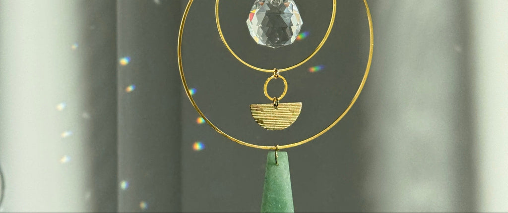 Vertical gold geometric suncatcher with three tiered hoops, a faceted crystal sphere, and two green Aventurine stones casting a galaxy of rainbows on a white wall.