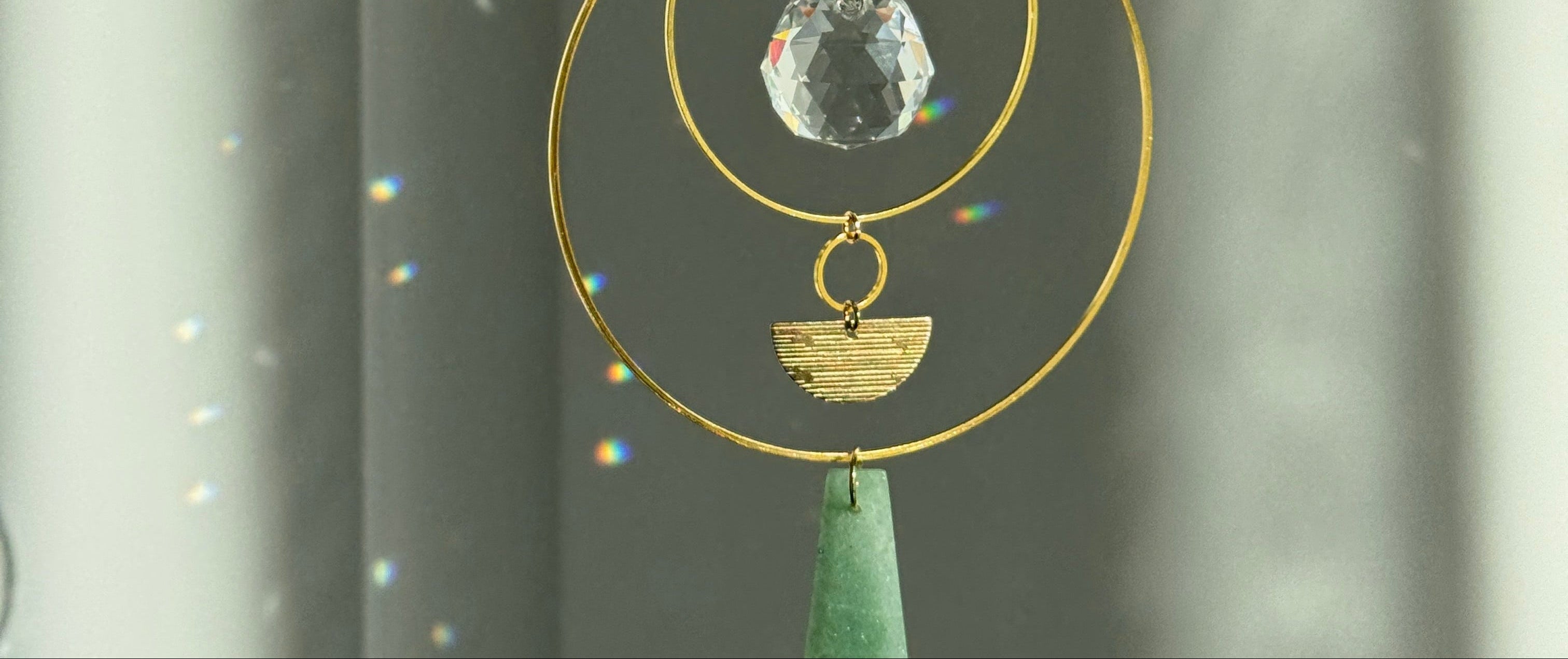 Vertical gold geometric suncatcher with three tiered hoops, a faceted crystal sphere, and two green Aventurine stones casting a galaxy of rainbows on a white wall.