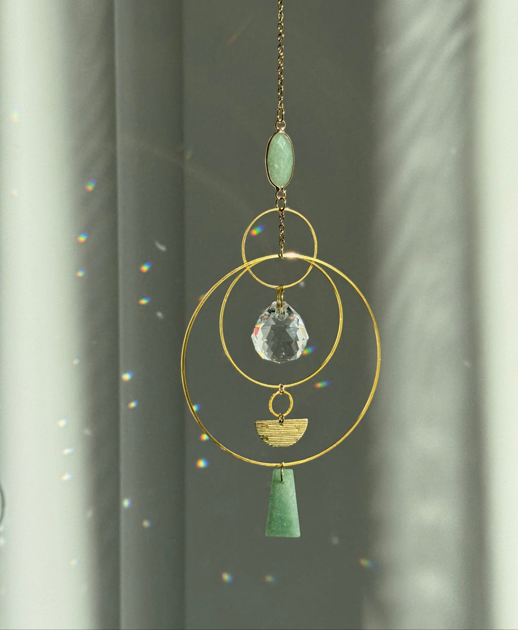 Decorative hanging ornament with gold chain, green stones, and crystal-like center against a neutral background