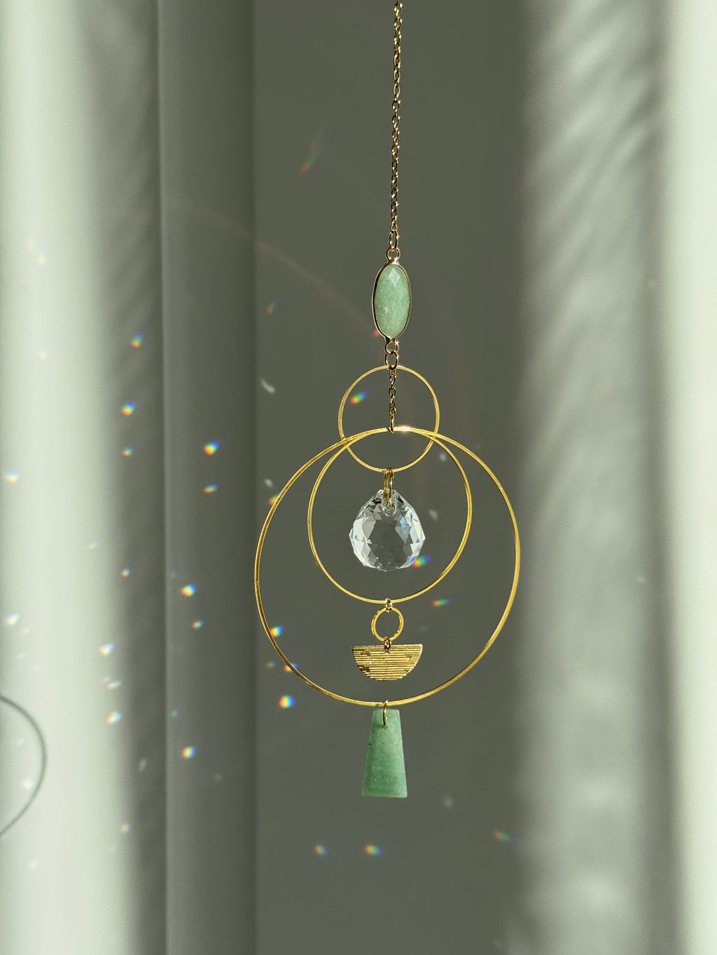 Vertical gold geometric suncatcher with three tiered hoops, a faceted crystal sphere, and two green Aventurine stones casting a galaxy of rainbows on a white wall.
