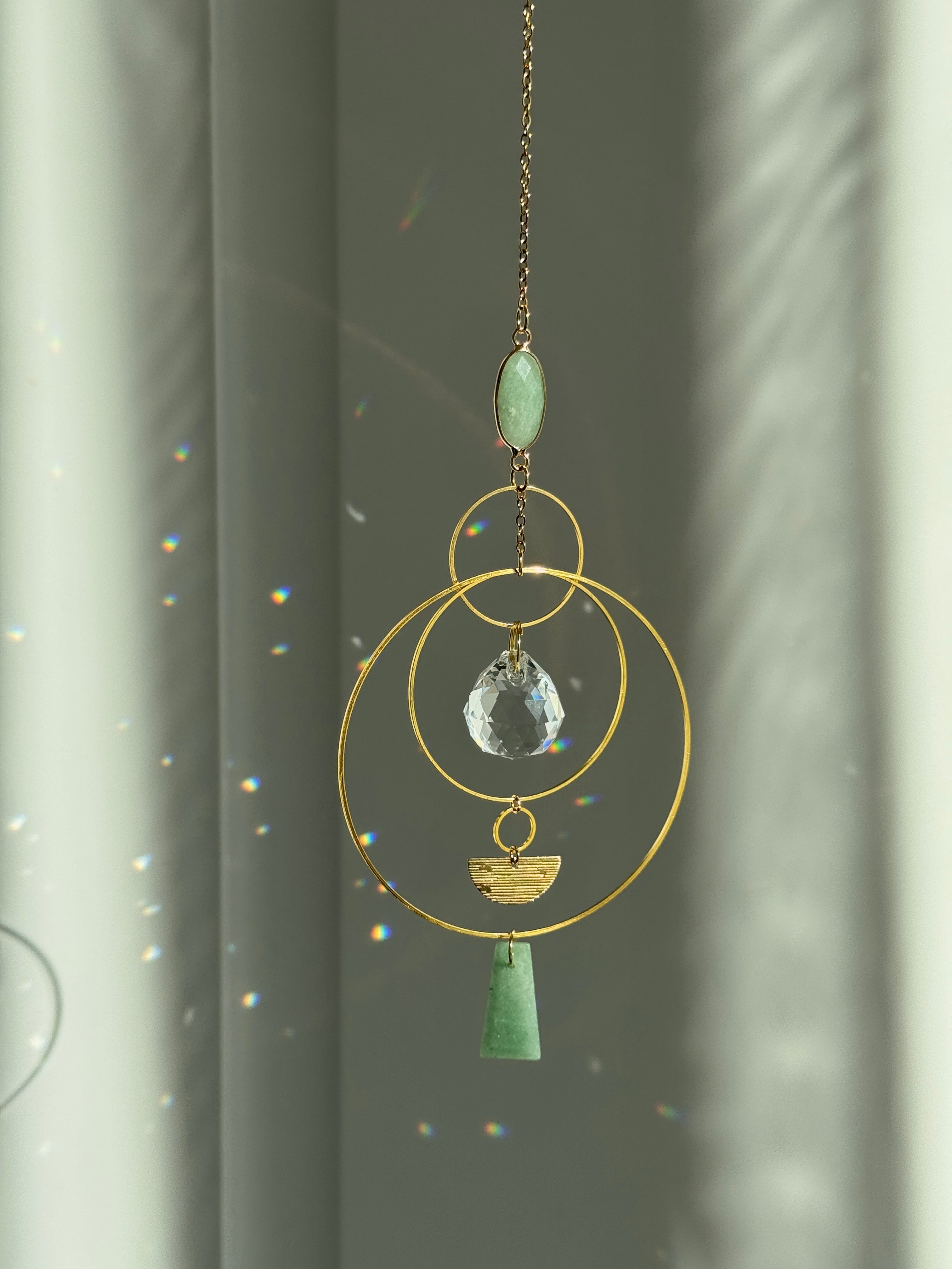 Vertical gold geometric suncatcher with three tiered hoops, a faceted crystal sphere, and two green Aventurine stones casting a galaxy of rainbows on a white wall.