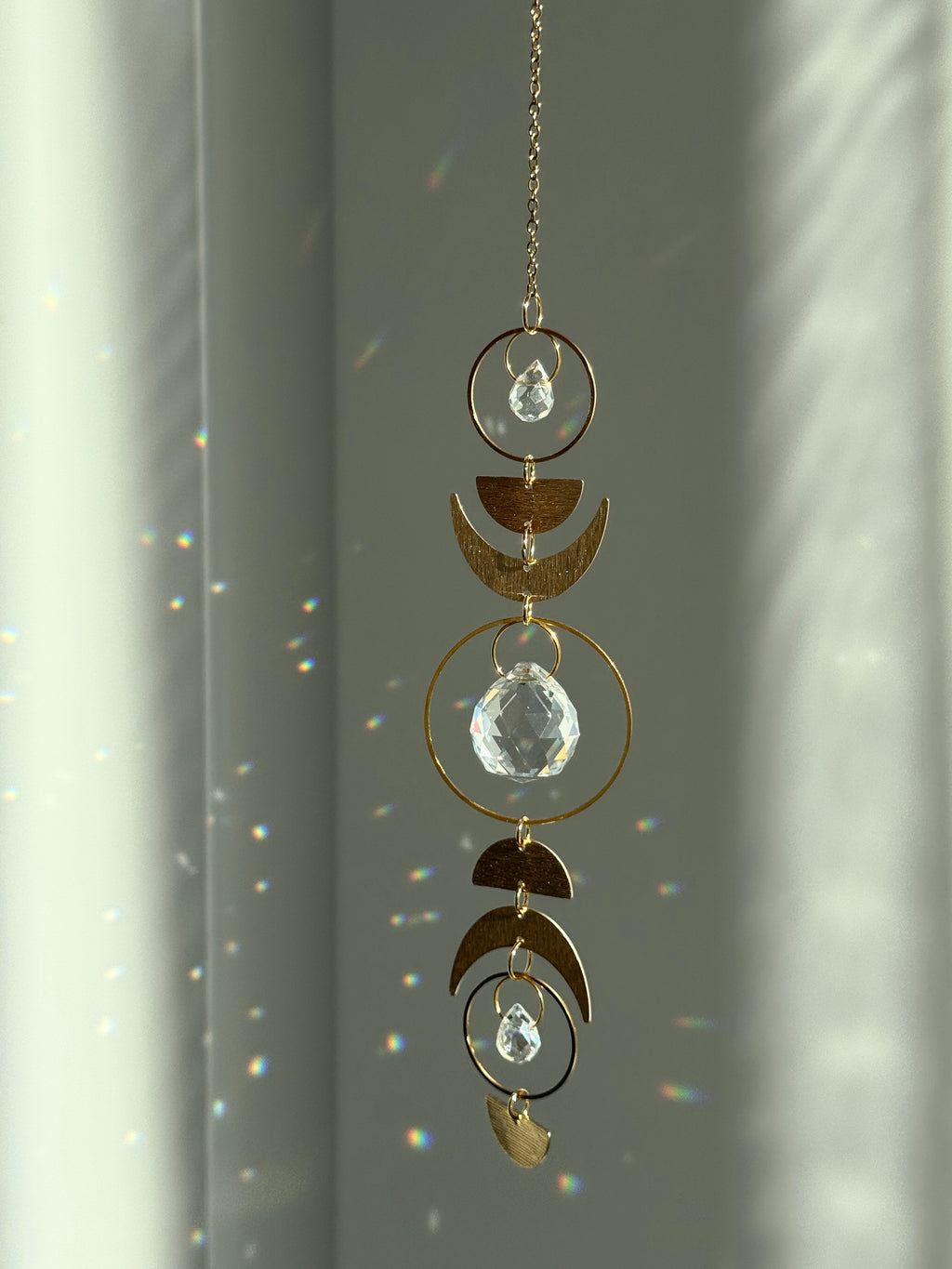 Handcrafted gold moon phase suncatcher featuring a vertical alignment of textured brass crescent moons and hoops with three clear crystal prisms casting rainbows on a white wall.