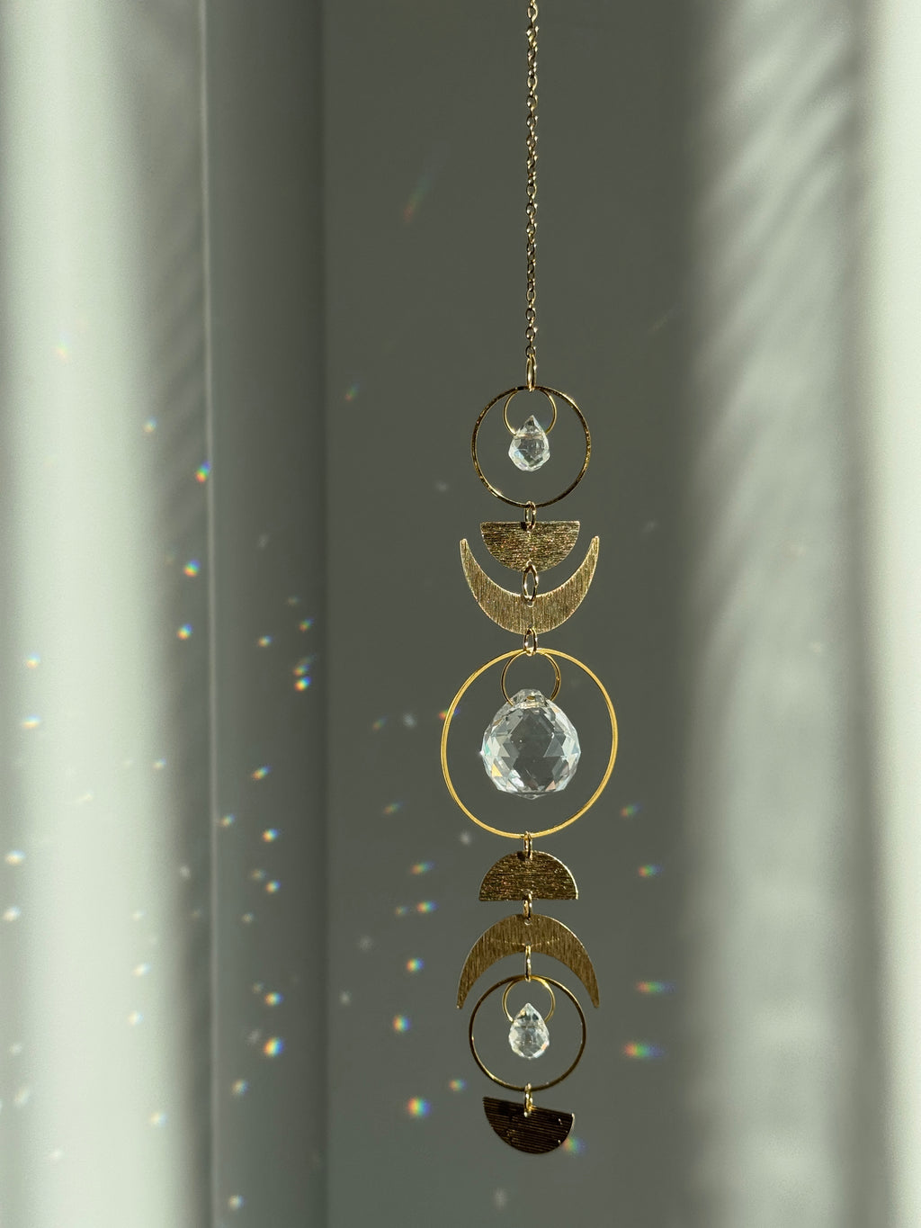 Handcrafted gold moon phase suncatcher featuring a vertical alignment of textured brass crescent moons and hoops with three clear crystal prisms casting rainbows on a white wall.