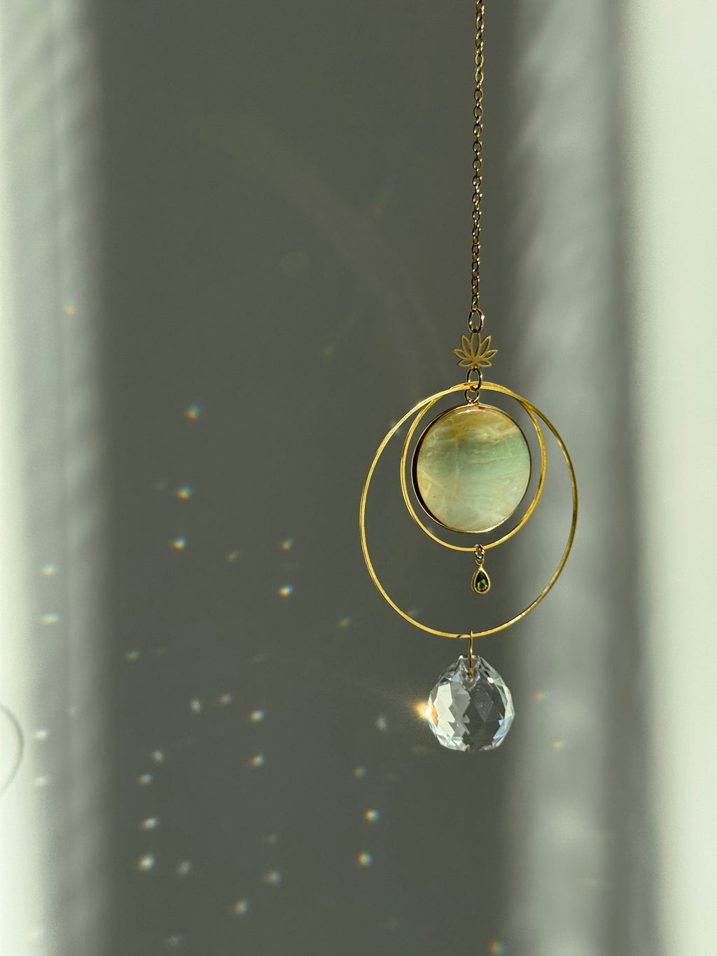 Detailed view of a gold lotus suncatcher featuring a round green Amazonite stone disc and a large faceted crystal drop reflecting rainbow light spots on a soft grey background.