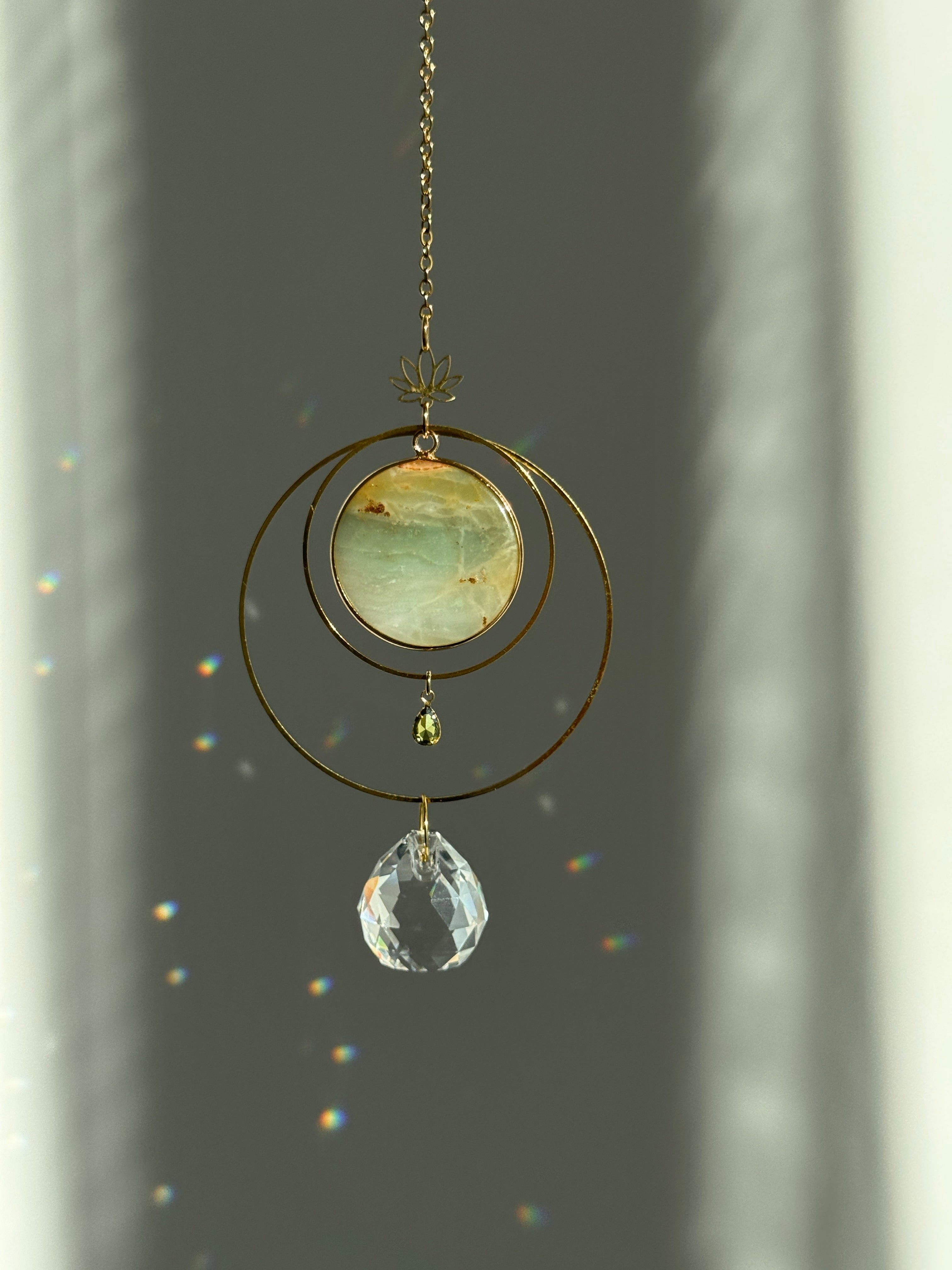 Detailed view of a gold lotus suncatcher featuring a round green Amazonite stone disc and a large faceted crystal drop reflecting rainbow light spots on a soft grey background.