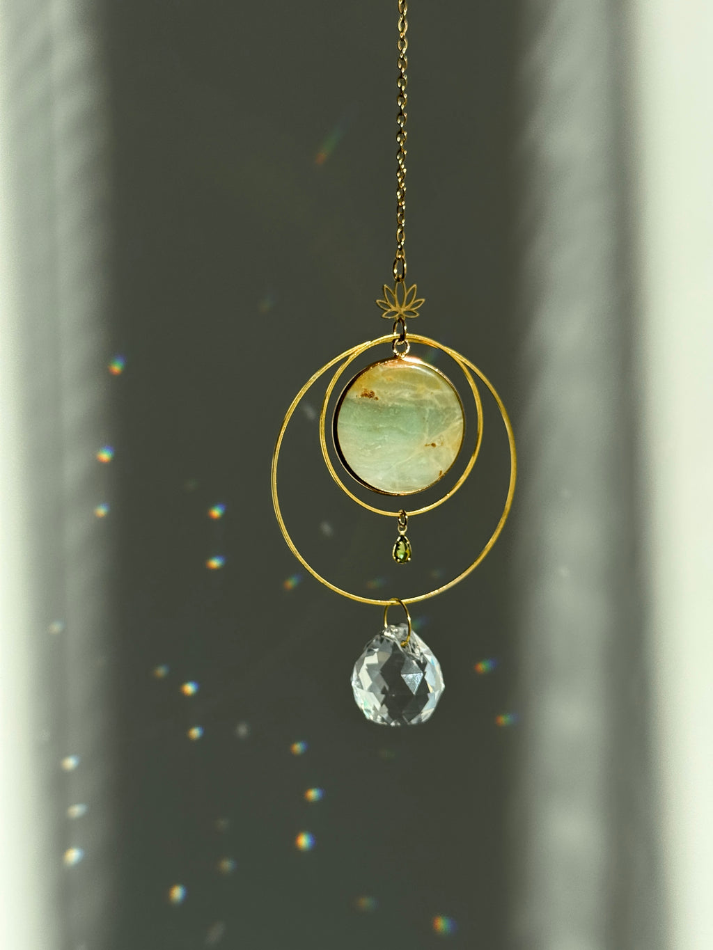 Detailed view of a gold lotus suncatcher featuring a round green Amazonite stone disc and a large faceted crystal drop reflecting rainbow light spots on a soft grey background.