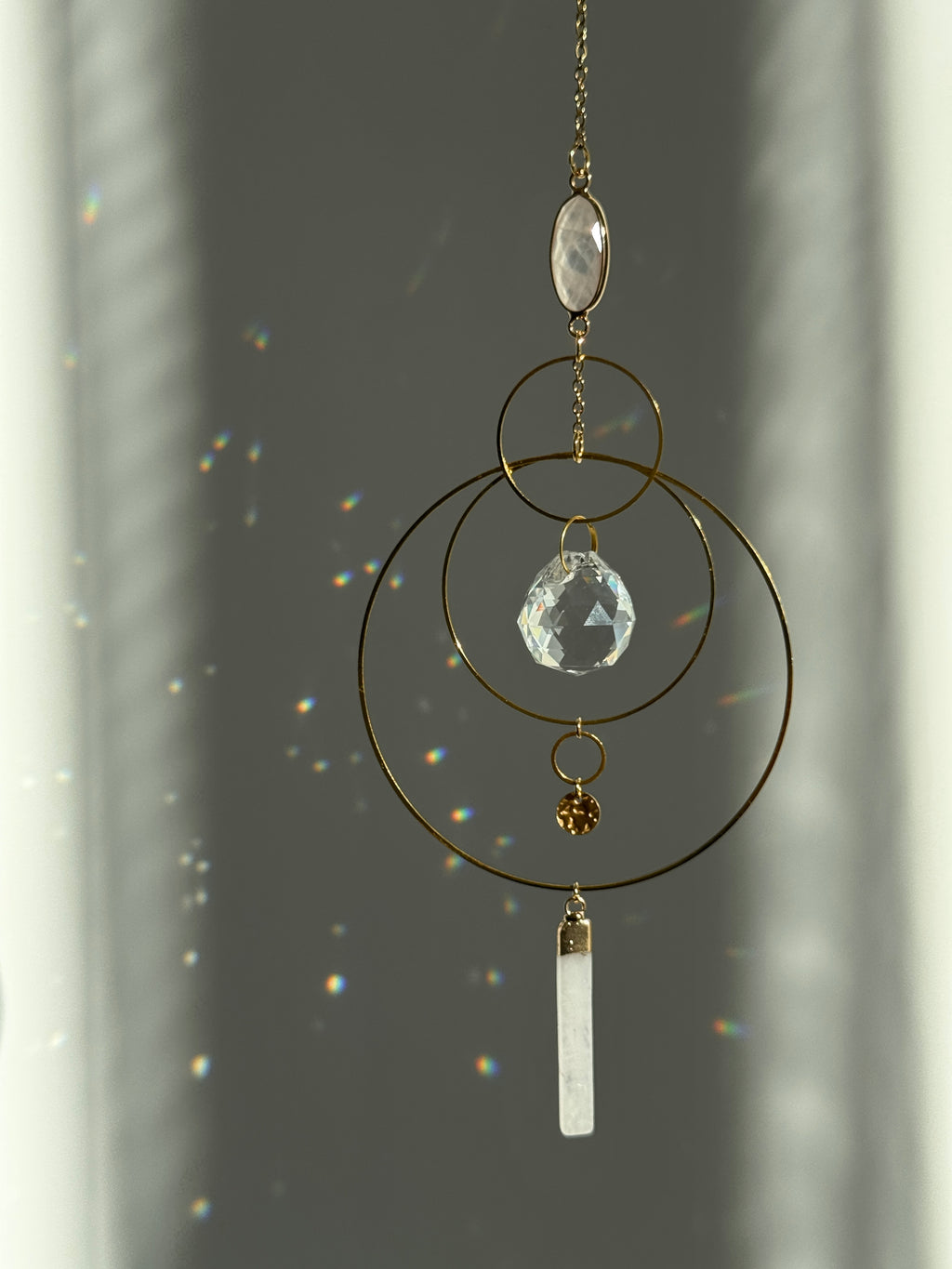 A gold geometric suncatcher featuring two pink Rose Quartz gemstones and a faceted crystal sphere. The suncatcher consists of two interlocking gold hoops, a textured coin charm, and is hanging against a soft-focus background, reflecting bright light.