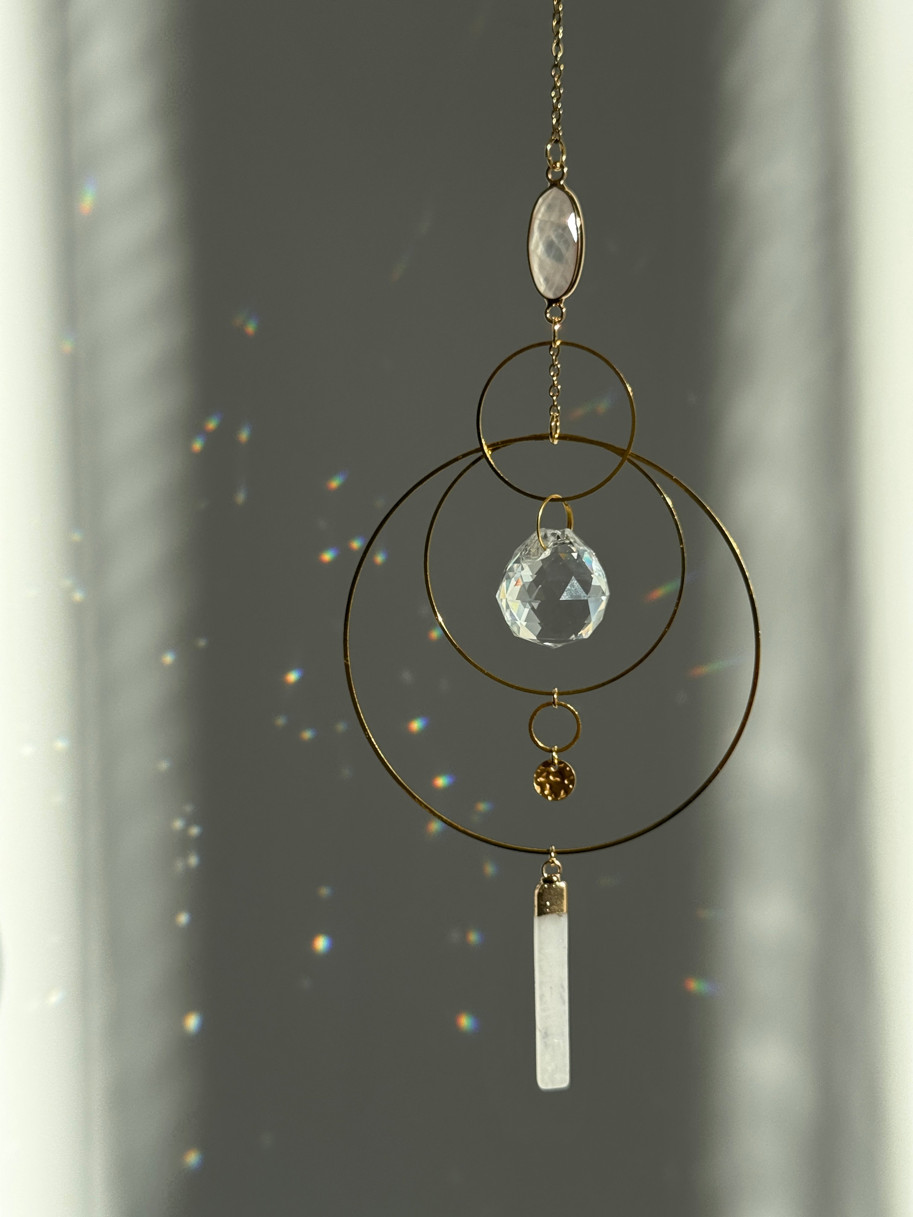 A gold geometric suncatcher featuring two pink Rose Quartz gemstones and a faceted crystal sphere. The suncatcher consists of two interlocking gold hoops, a textured coin charm, and is hanging against a soft-focus background, reflecting bright light.