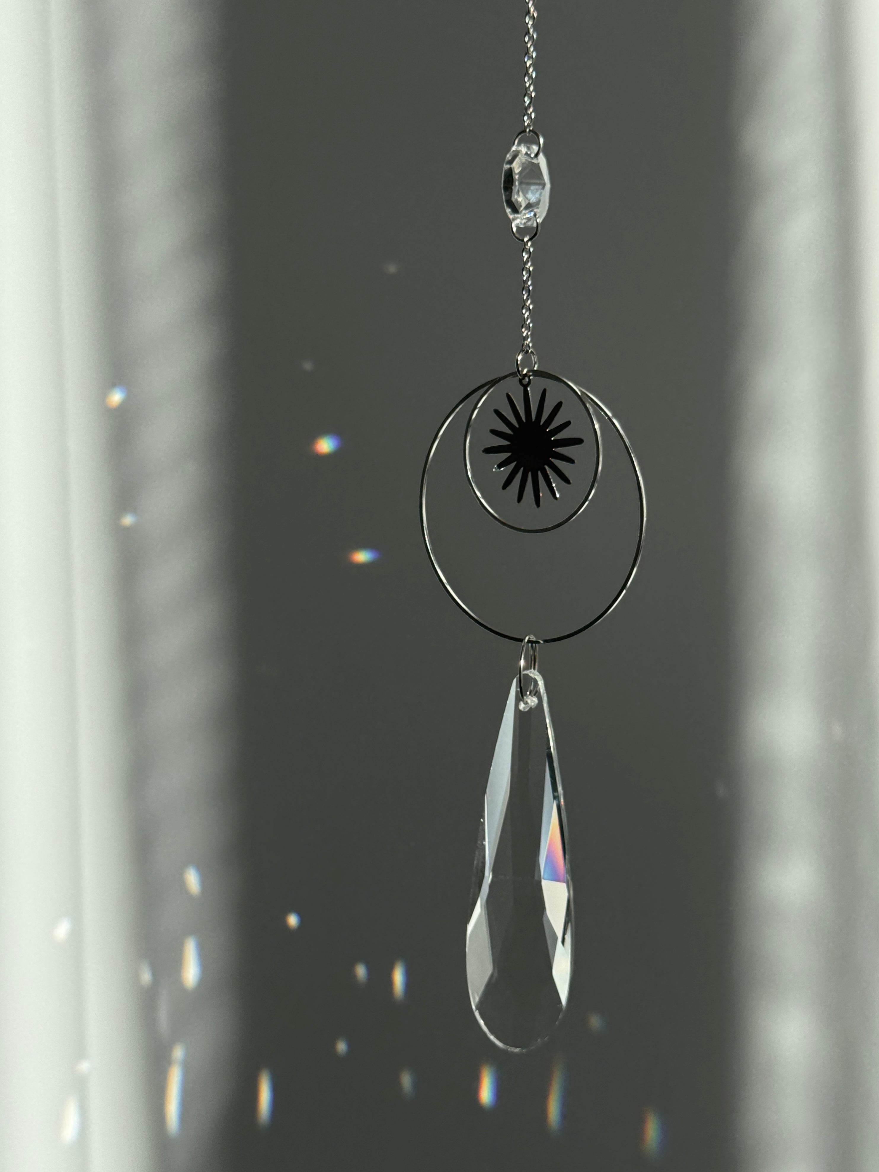 Handmade silver suncatcher with a minimalist starburst design and a large faceted teardrop crystal prism, hanging in natural sunlight with rainbow reflections and shadows on a grey wall background.