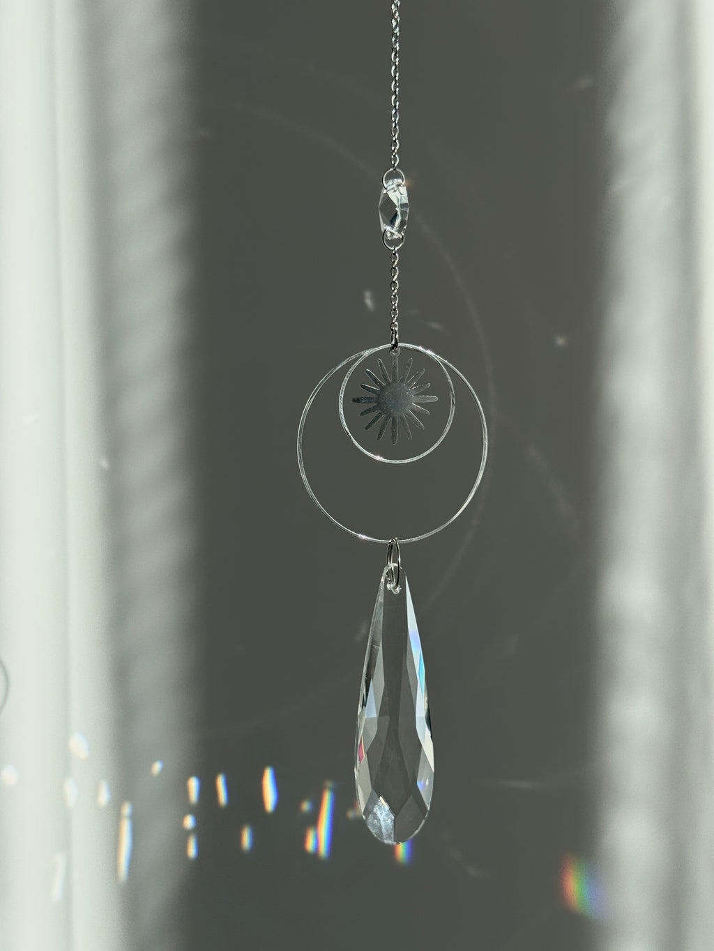 Handmade silver suncatcher with a minimalist starburst design and a large faceted teardrop crystal prism, hanging in natural sunlight with rainbow reflections and shadows on a grey wall background.