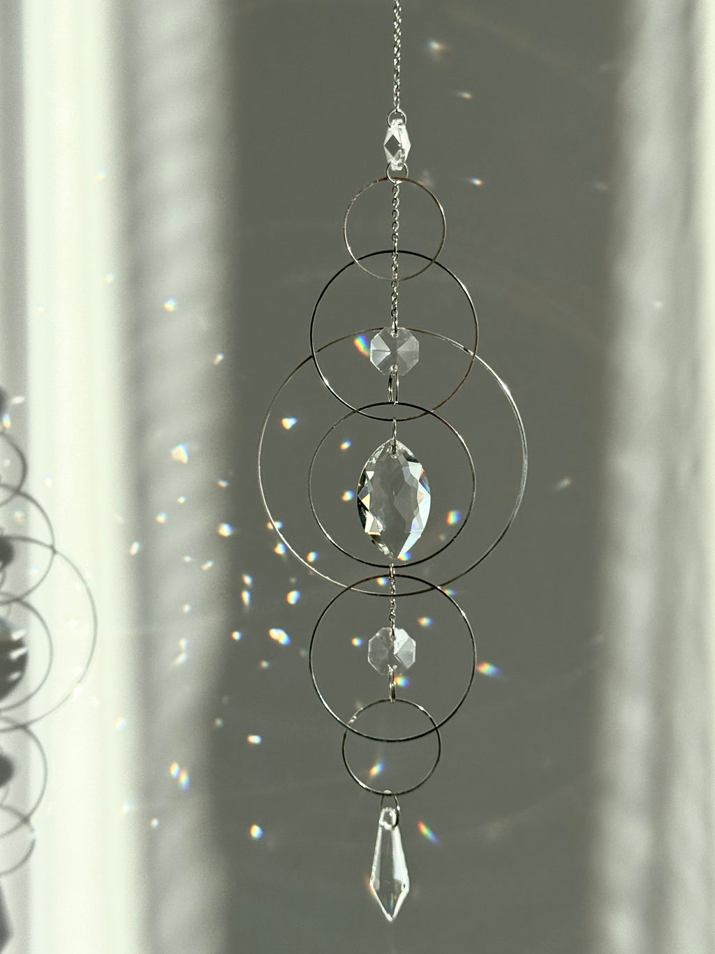 60cm silver geometric suncatcher featuring interlocking concentric hoops and clear marquise-cut crystal prisms, hanging in a window and casting rainbows on a wall.