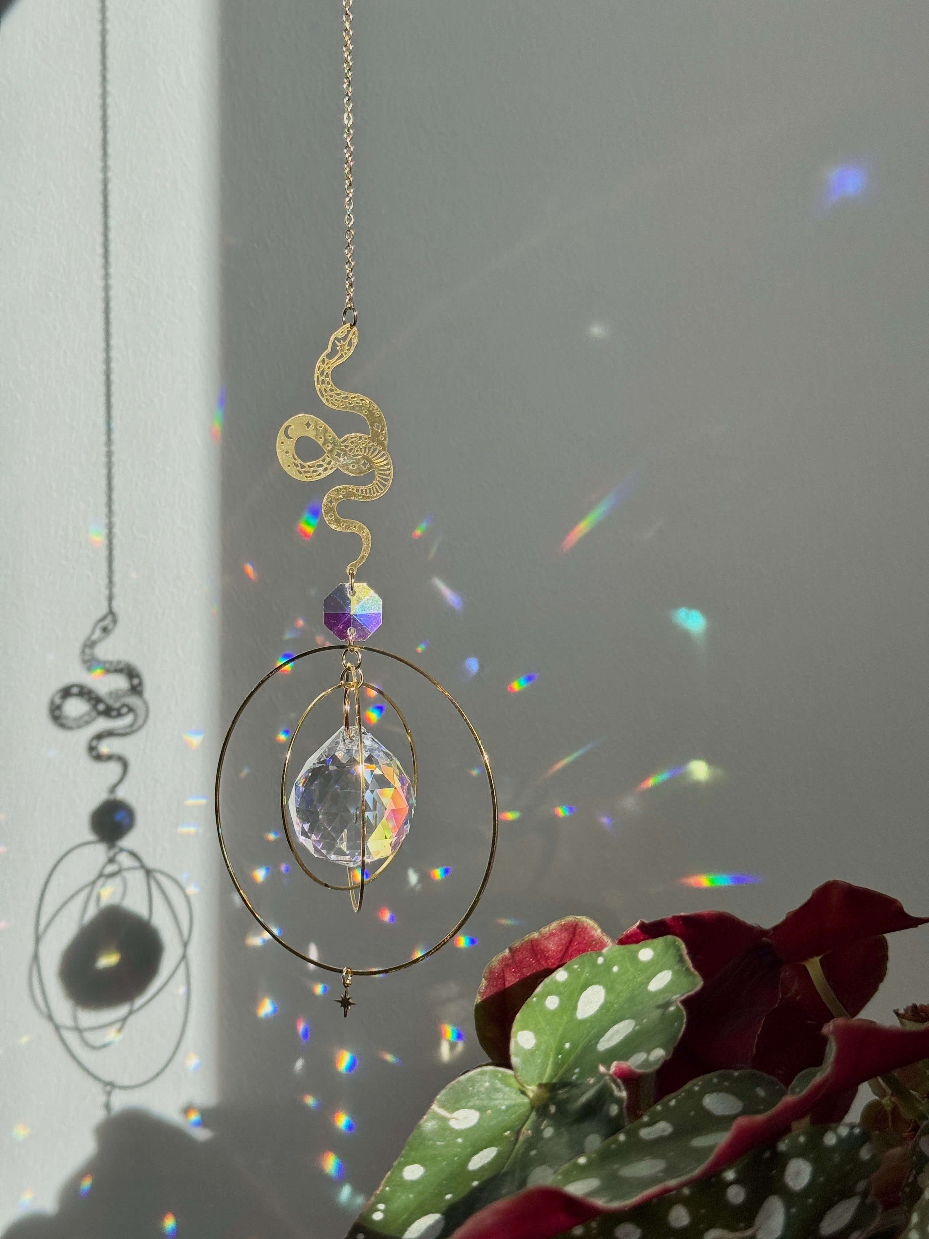 Handcrafted Sunkissed Heartbeat suncatcher with a faceted crystal heart and gold orbital rings casting vibrant rainbows on a white wall near a green plant.