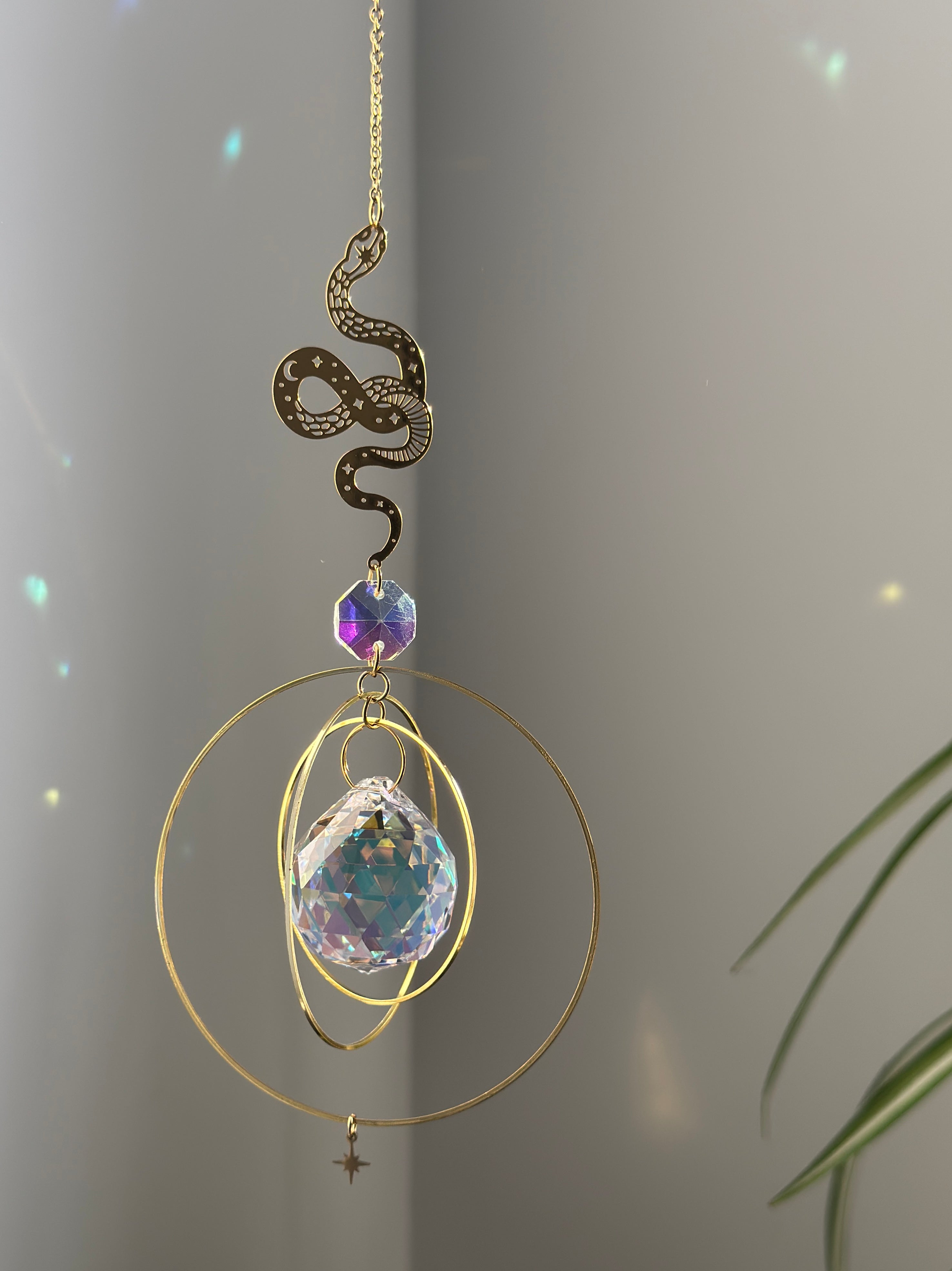 Handcrafted Sunkissed Heartbeat suncatcher with a faceted crystal heart and gold orbital rings casting vibrant rainbows on a white wall near a green plant.
