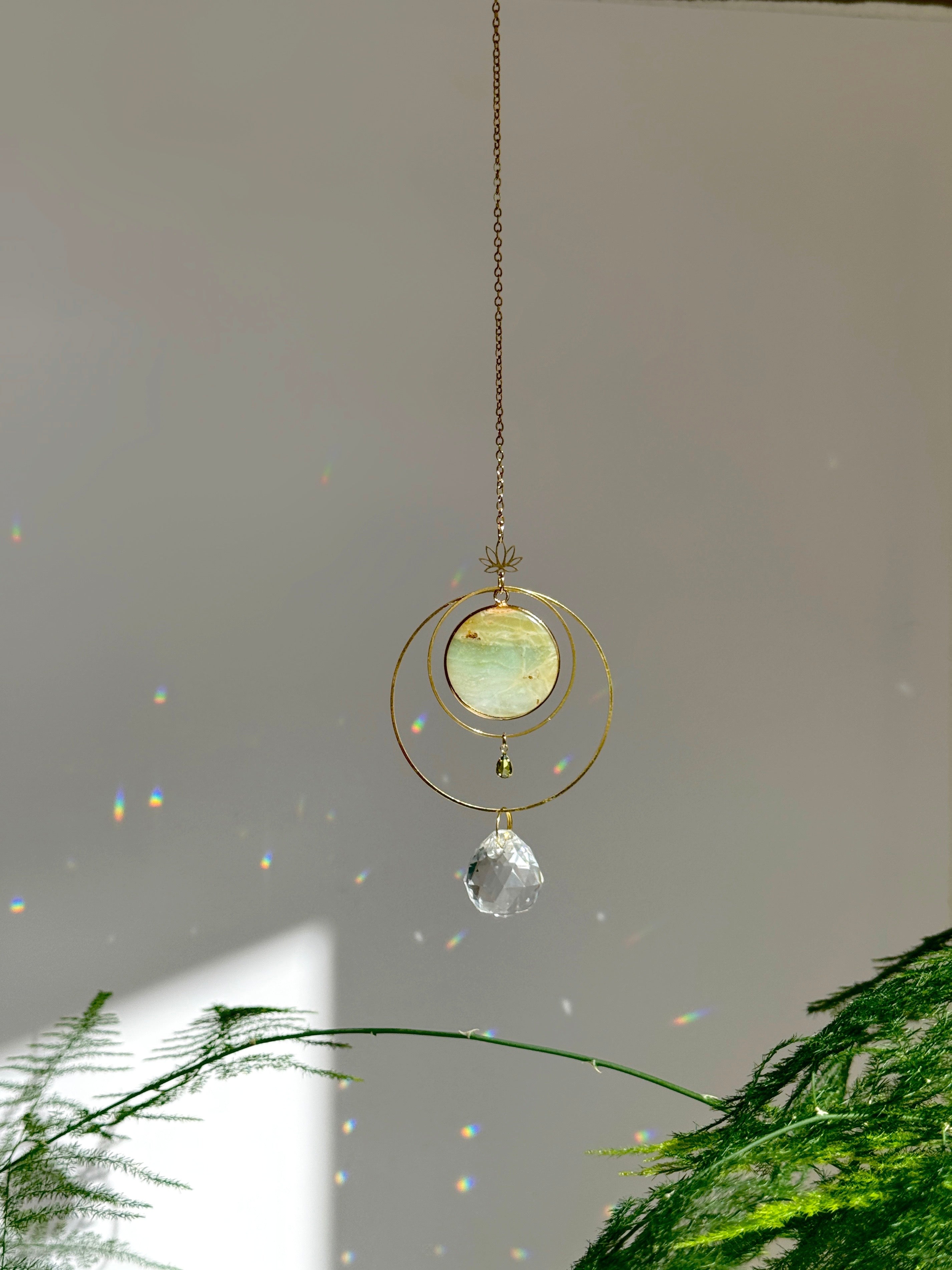 Detailed view of a gold lotus suncatcher featuring a round green Amazonite stone disc and a large faceted crystal drop reflecting rainbow light spots on a soft grey background.