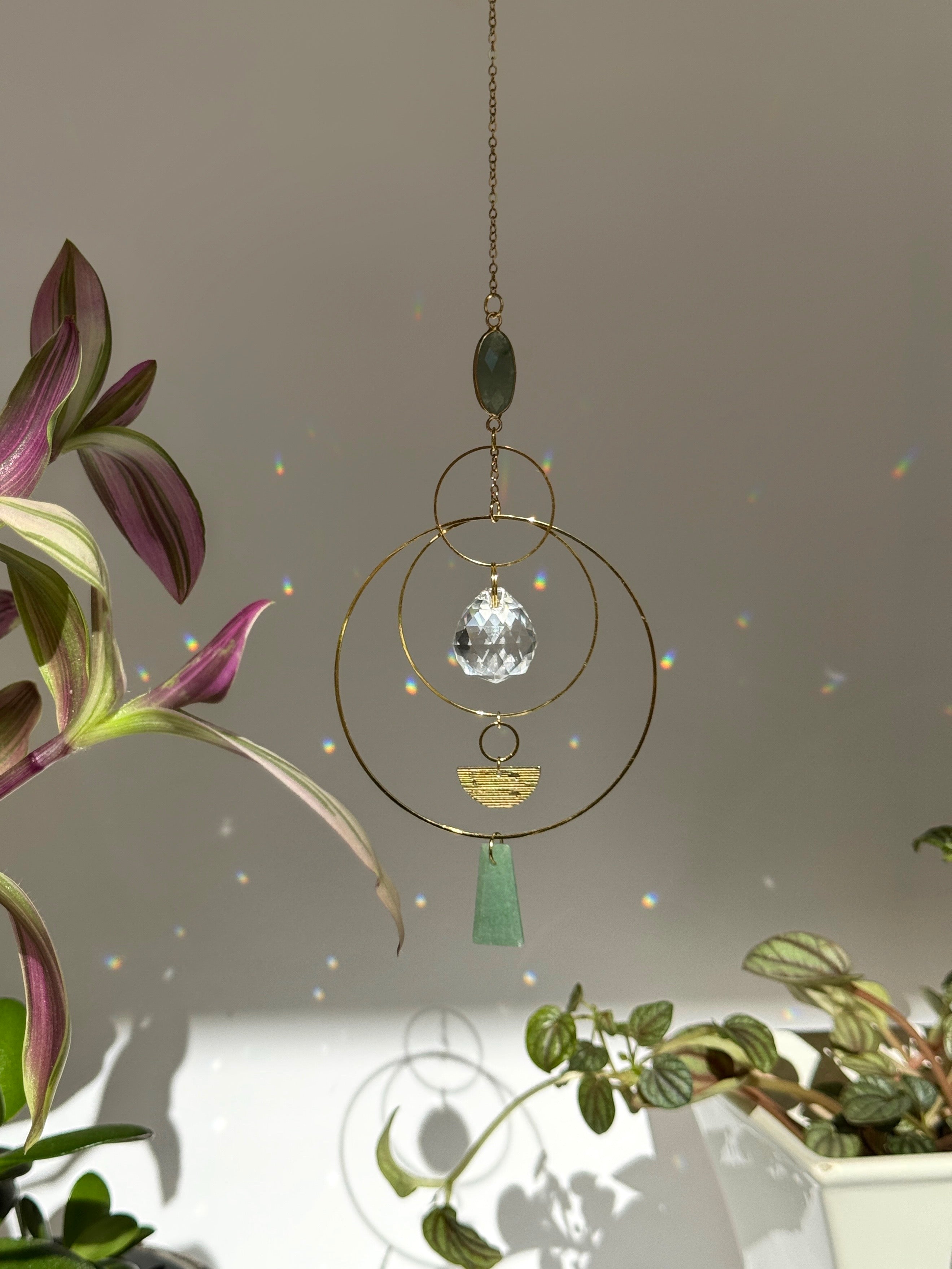 Vertical gold geometric suncatcher with three tiered hoops, a faceted crystal sphere, and two green Aventurine stones casting a galaxy of rainbows on a white wall.