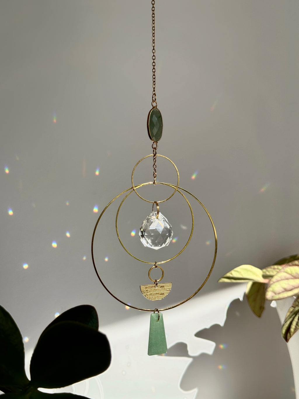 Vertical gold geometric suncatcher with three tiered hoops, a faceted crystal sphere, and two green Aventurine stones casting a galaxy of rainbows on a white wall.