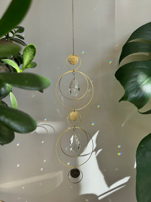Symmetry in Light – Handmade geometric suncatcher with stainless steel rings and faceted crystal droplets. Celestial window hanging casting rainbows on a wall.