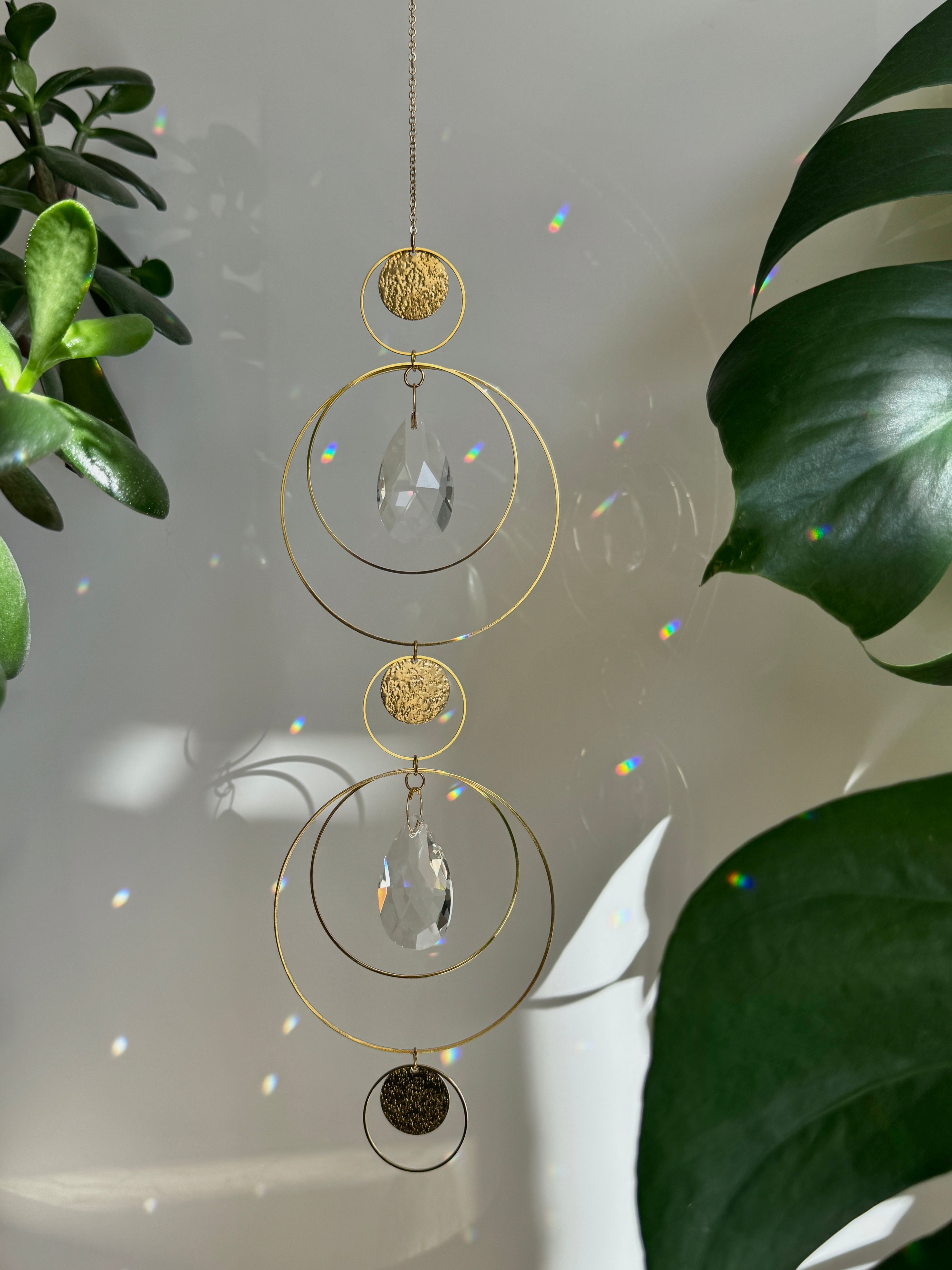 Symmetry in Light – Handmade geometric suncatcher with stainless steel rings and faceted crystal droplets. Celestial window hanging casting rainbows on a wall.