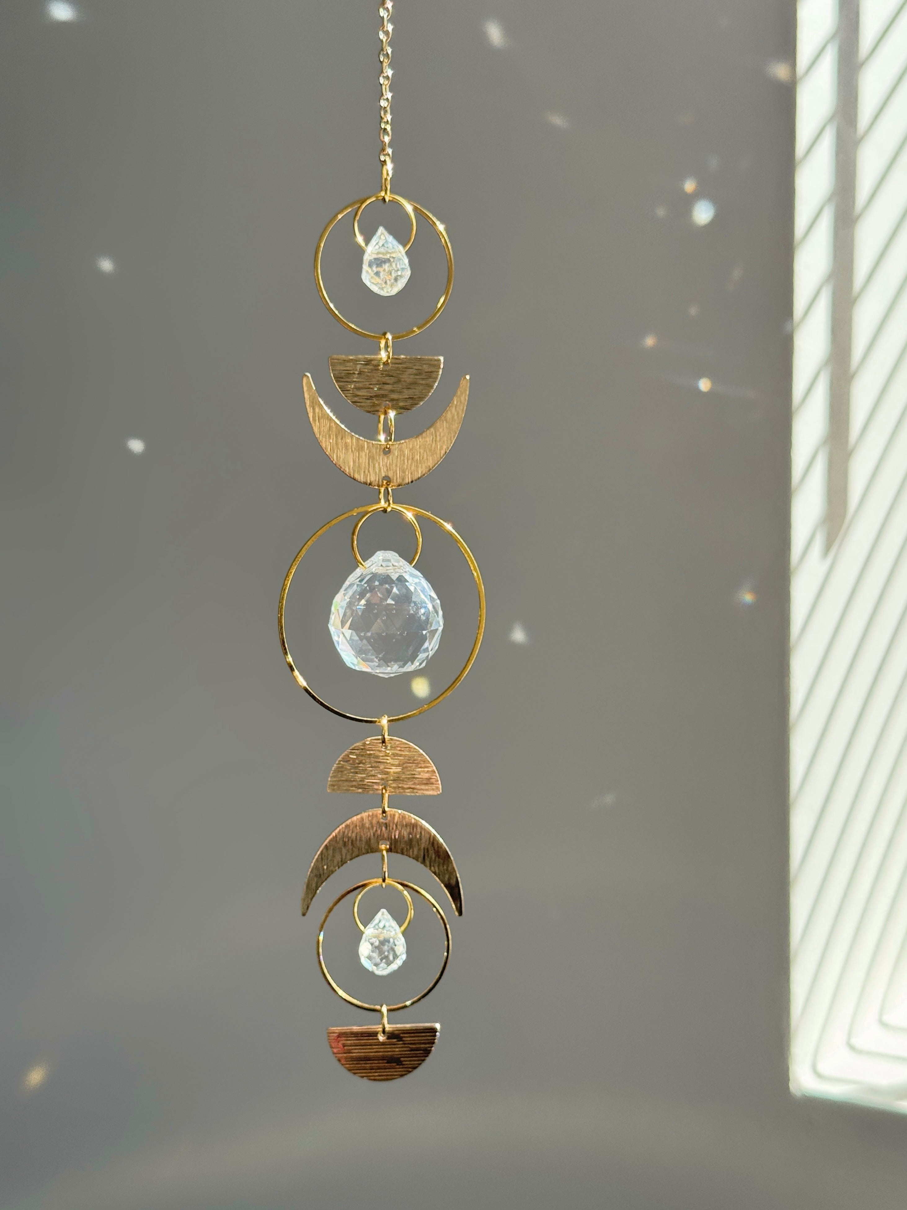 Handcrafted gold moon phase suncatcher featuring a vertical alignment of textured brass crescent moons and hoops with three clear crystal prisms casting rainbows on a white wall.