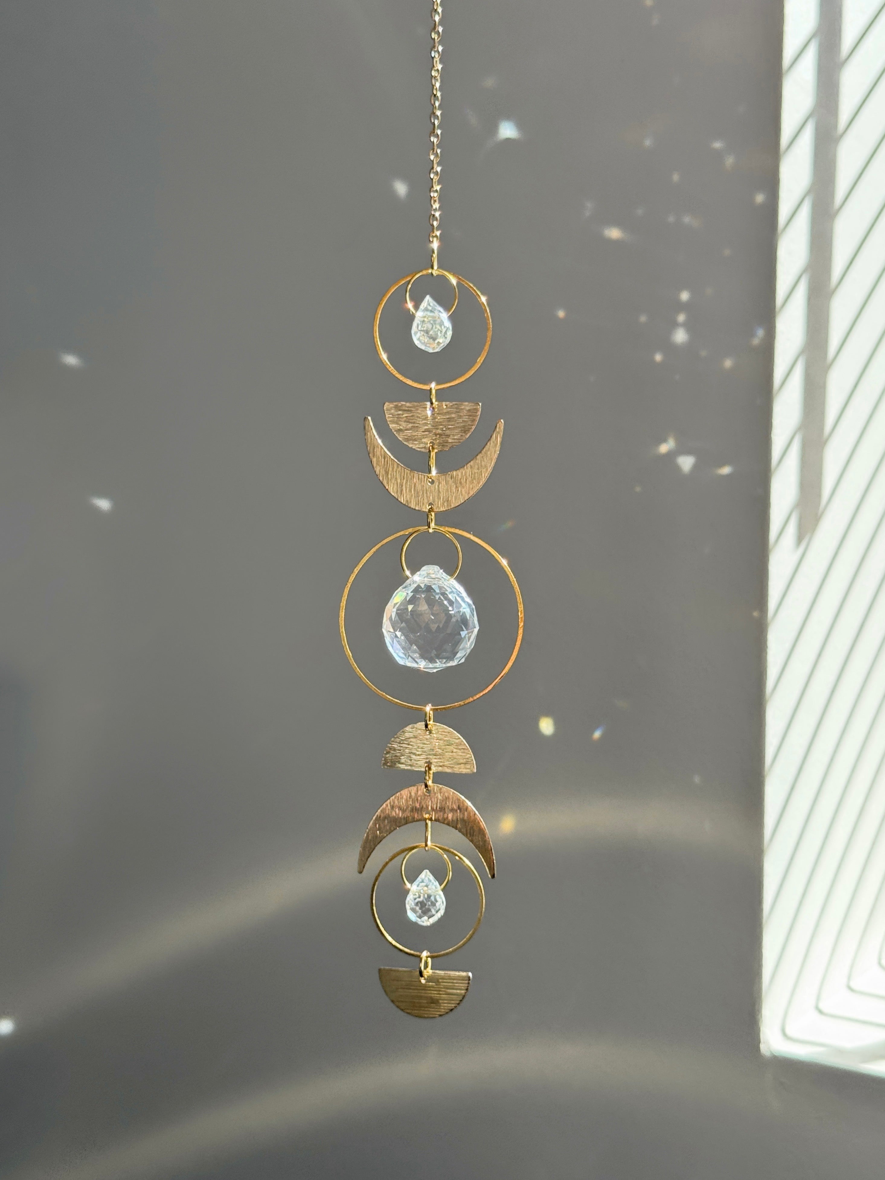 Handcrafted gold moon phase suncatcher featuring a vertical alignment of textured brass crescent moons and hoops with three clear crystal prisms casting rainbows on a white wall.
