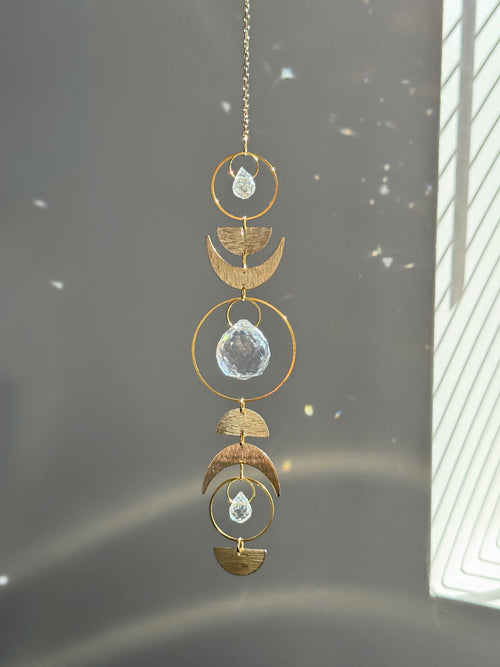 Handcrafted gold moon phase suncatcher featuring a vertical alignment of textured brass crescent moons and hoops with three clear crystal prisms casting rainbows on a white wall.