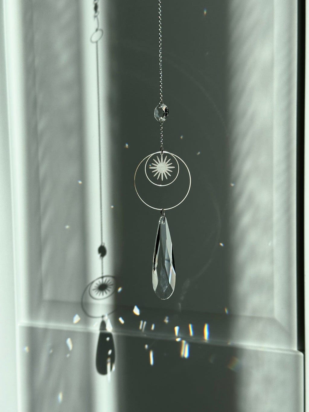 Handmade silver suncatcher with a minimalist starburst design and a large faceted teardrop crystal prism, hanging in natural sunlight with rainbow reflections and shadows on a grey wall background.
