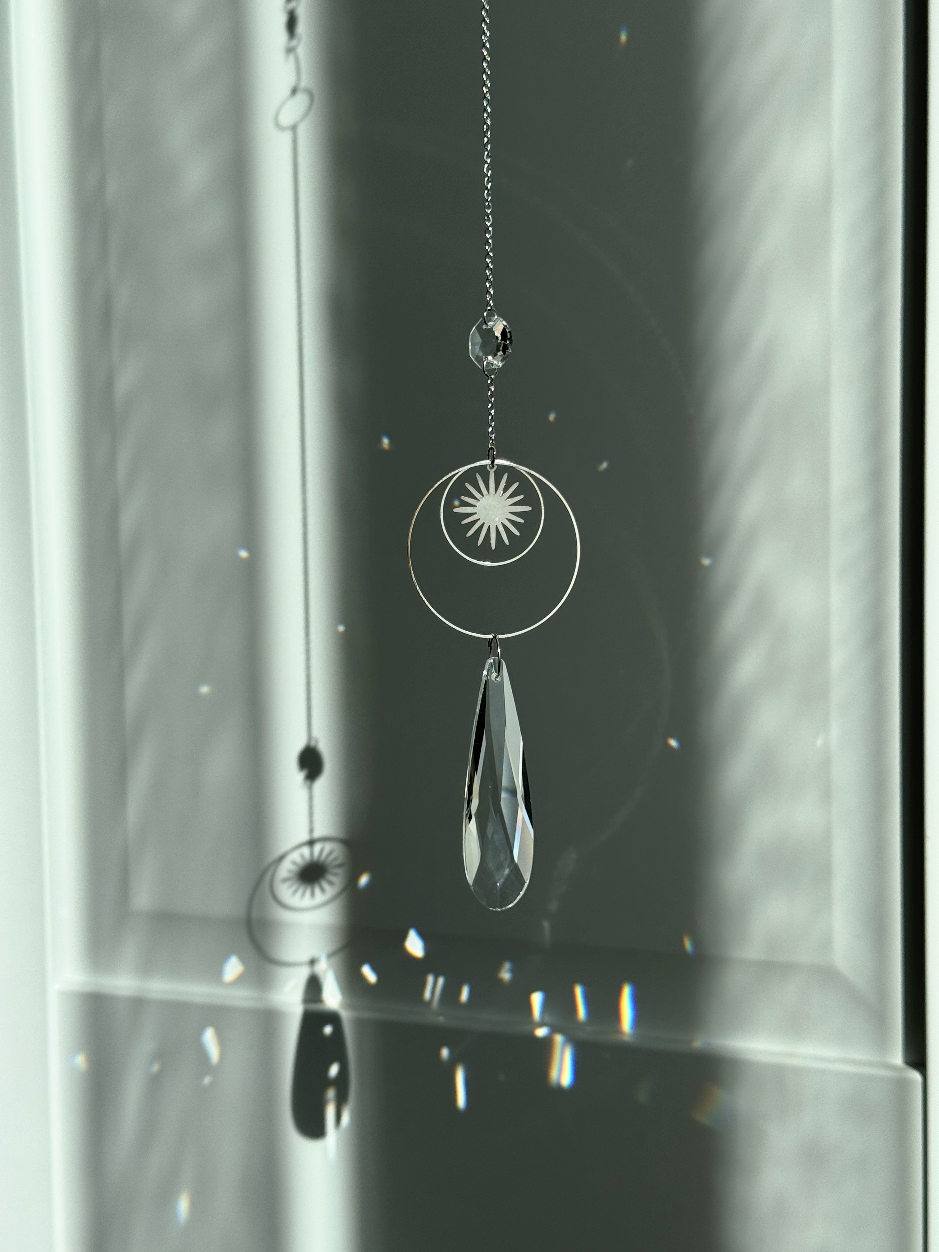 Handmade silver suncatcher with a minimalist starburst design and a large faceted teardrop crystal prism, hanging in natural sunlight with rainbow reflections and shadows on a grey wall background.