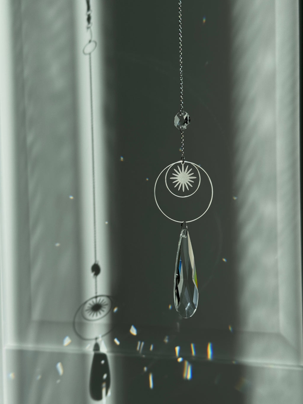 Handmade silver suncatcher with a minimalist starburst design and a large faceted teardrop crystal prism, hanging in natural sunlight with rainbow reflections and shadows on a grey wall background.