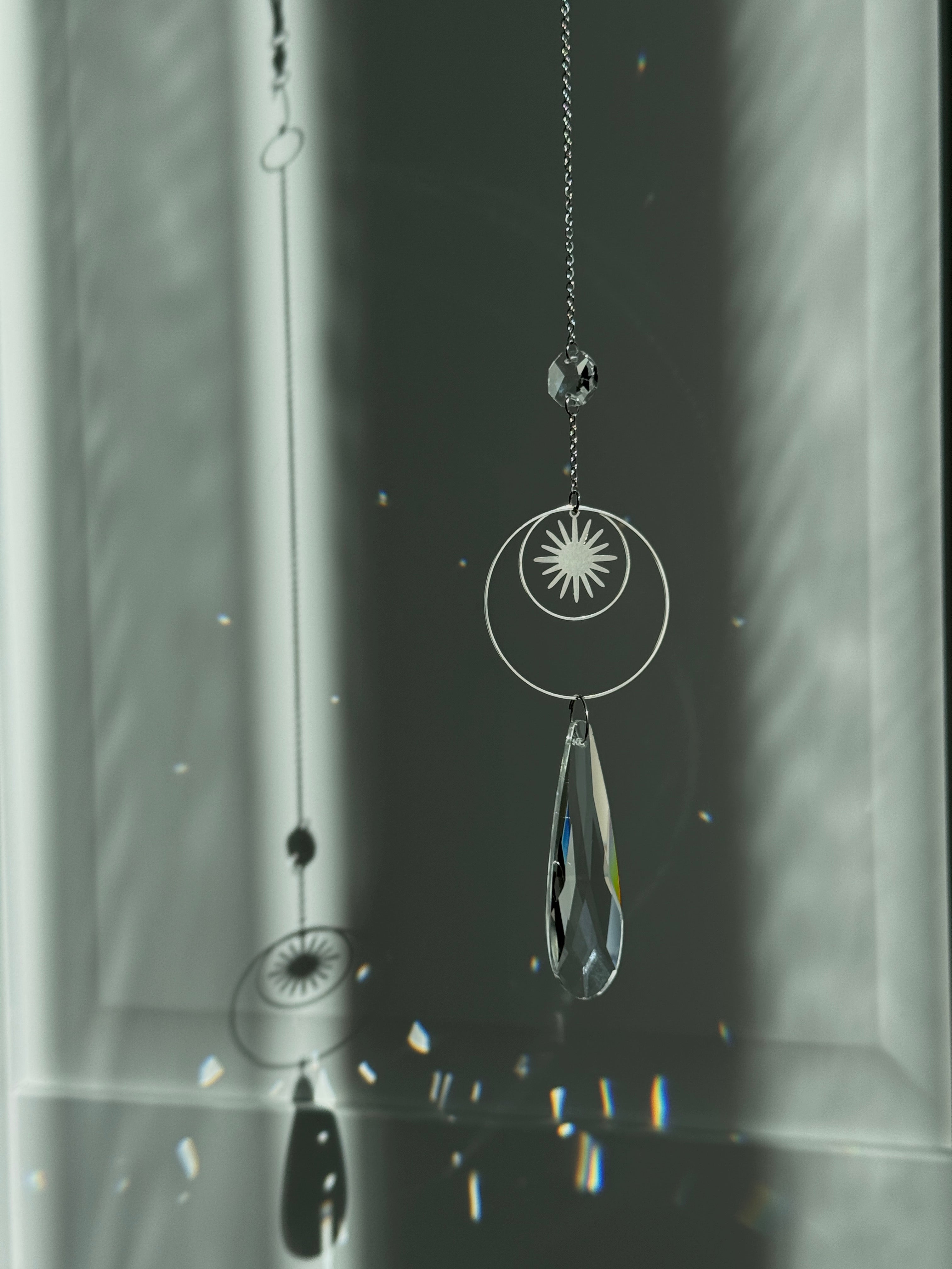 Handmade silver suncatcher with a minimalist starburst design and a large faceted teardrop crystal prism, hanging in natural sunlight with rainbow reflections and shadows on a grey wall background.