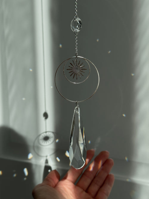 Handmade silver suncatcher with a minimalist starburst design and a large faceted teardrop crystal prism, hanging in natural sunlight with rainbow reflections and shadows on a grey wall background.