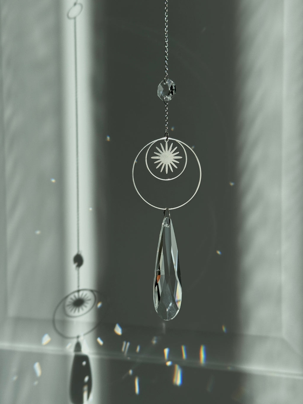 Handmade silver suncatcher with a minimalist starburst design and a large faceted teardrop crystal prism, hanging in natural sunlight with rainbow reflections and shadows on a grey wall background.