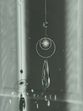 Handmade silver suncatcher with a minimalist starburst design and a large faceted teardrop crystal prism, hanging in natural sunlight with rainbow reflections and shadows on a grey wall background.