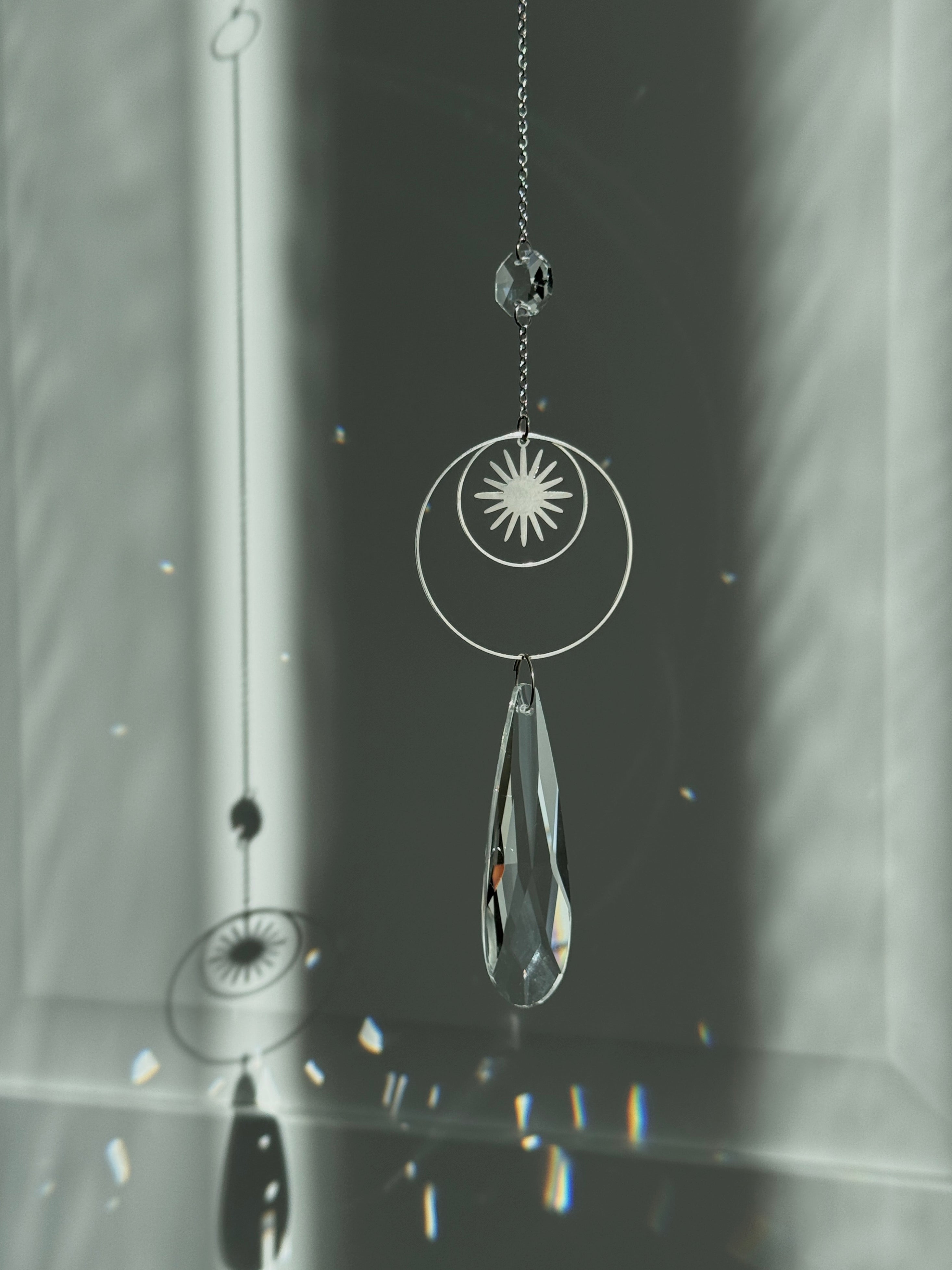 Handmade silver suncatcher with a minimalist starburst design and a large faceted teardrop crystal prism, hanging in natural sunlight with rainbow reflections and shadows on a grey wall background.