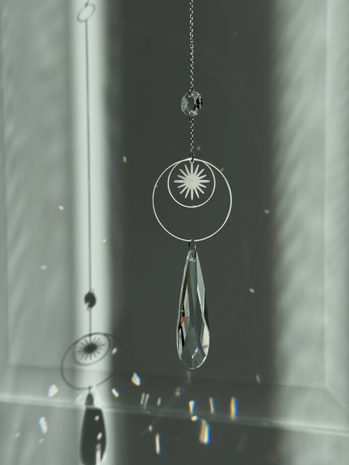 Handmade silver suncatcher with a minimalist starburst design and a large faceted teardrop crystal prism, hanging in natural sunlight with rainbow reflections and shadows on a grey wall background.