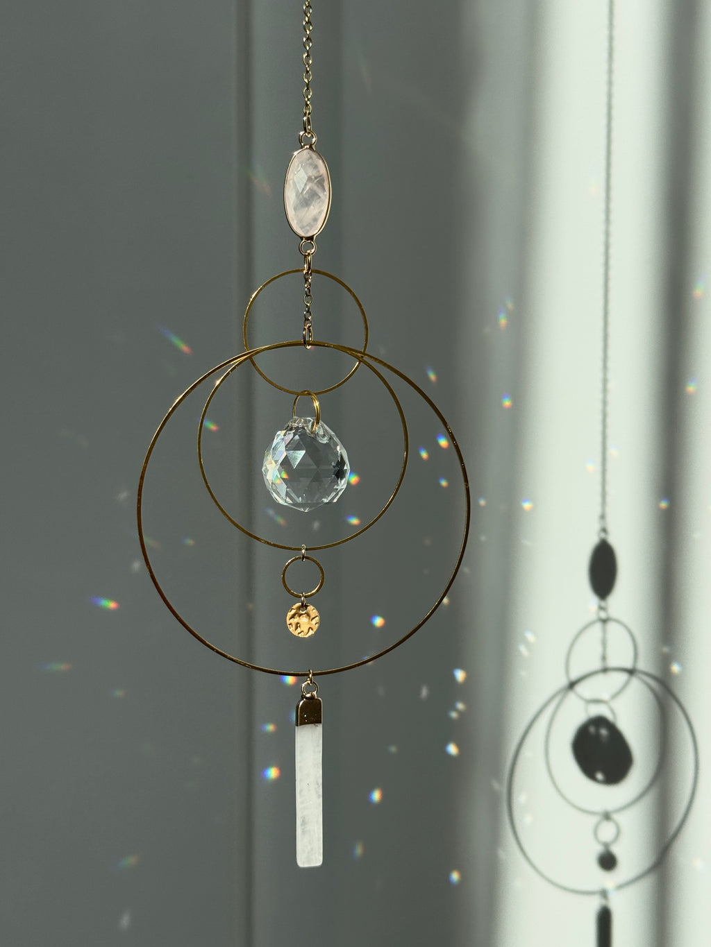 A gold geometric suncatcher featuring two pink Rose Quartz gemstones and a faceted crystal sphere. The suncatcher consists of two interlocking gold hoops, a textured coin charm, and is hanging against a soft-focus background, reflecting bright light.