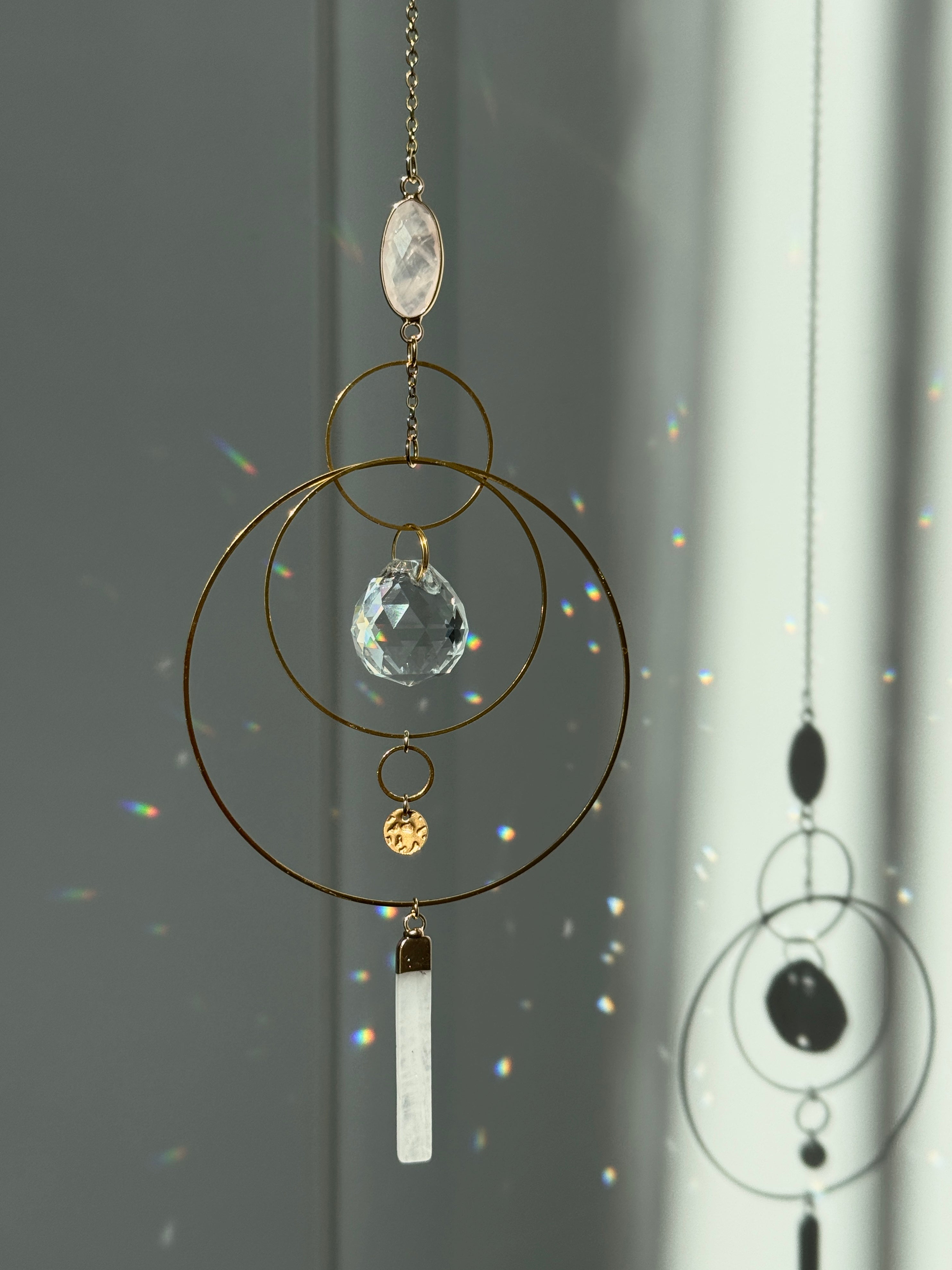 A gold geometric suncatcher featuring two pink Rose Quartz gemstones and a faceted crystal sphere. The suncatcher consists of two interlocking gold hoops, a textured coin charm, and is hanging against a soft-focus background, reflecting bright light.