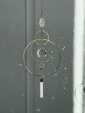 A gold geometric suncatcher featuring two pink Rose Quartz gemstones and a faceted crystal sphere. The suncatcher consists of two interlocking gold hoops, a textured coin charm, and is hanging against a soft-focus background, reflecting bright light.