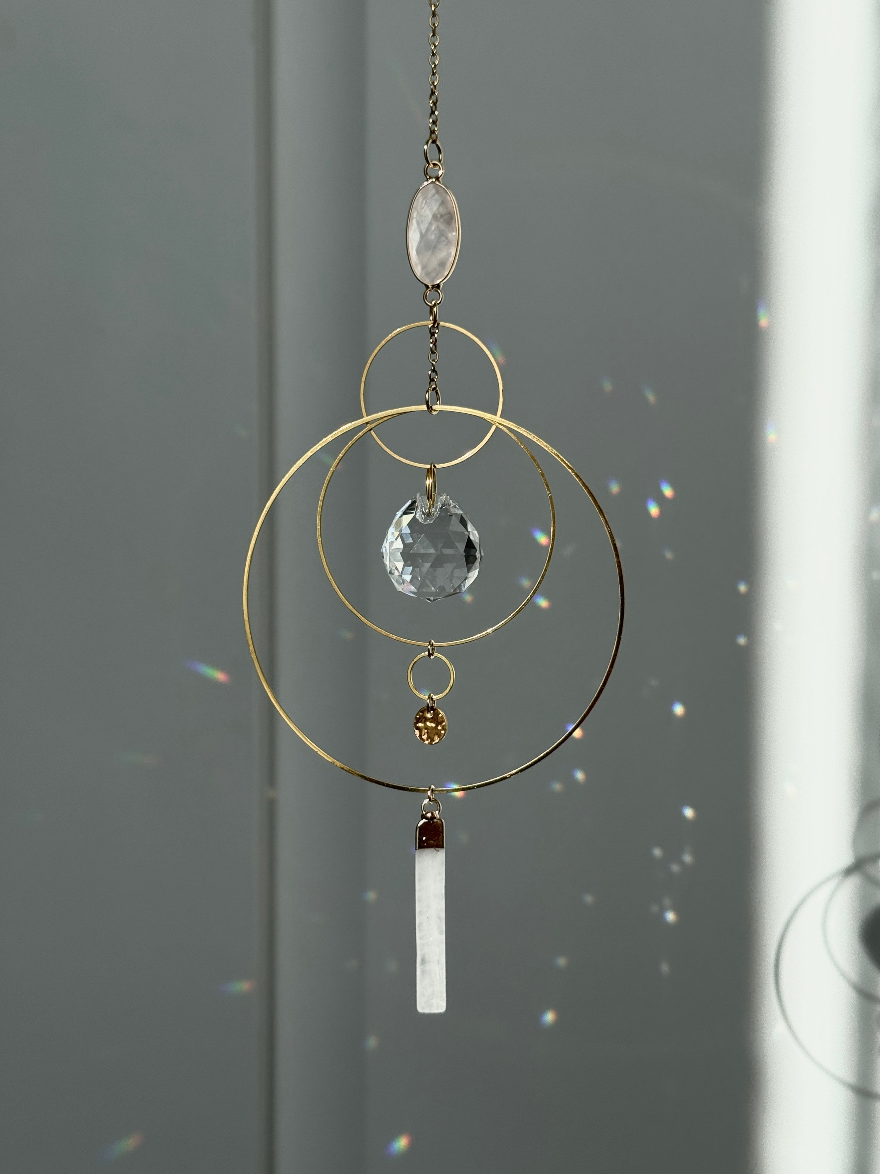 A gold geometric suncatcher featuring two pink Rose Quartz gemstones and a faceted crystal sphere. The suncatcher consists of two interlocking gold hoops, a textured coin charm, and is hanging against a soft-focus background, reflecting bright light.