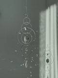 60cm silver geometric suncatcher featuring interlocking concentric hoops and clear marquise-cut crystal prisms, hanging in a window and casting rainbows on a wall.