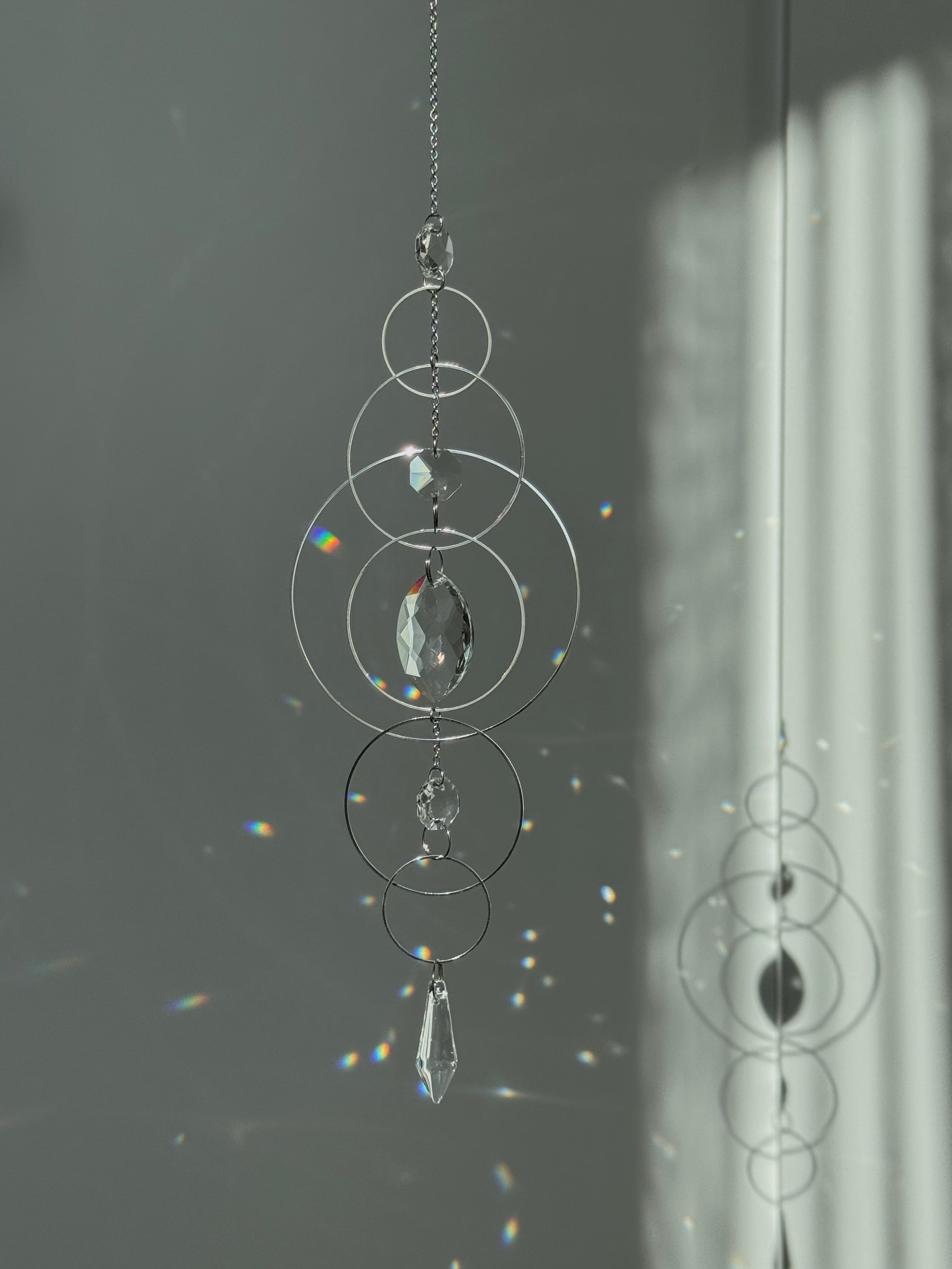 60cm silver geometric suncatcher featuring interlocking concentric hoops and clear marquise-cut crystal prisms, hanging in a window and casting rainbows on a wall.