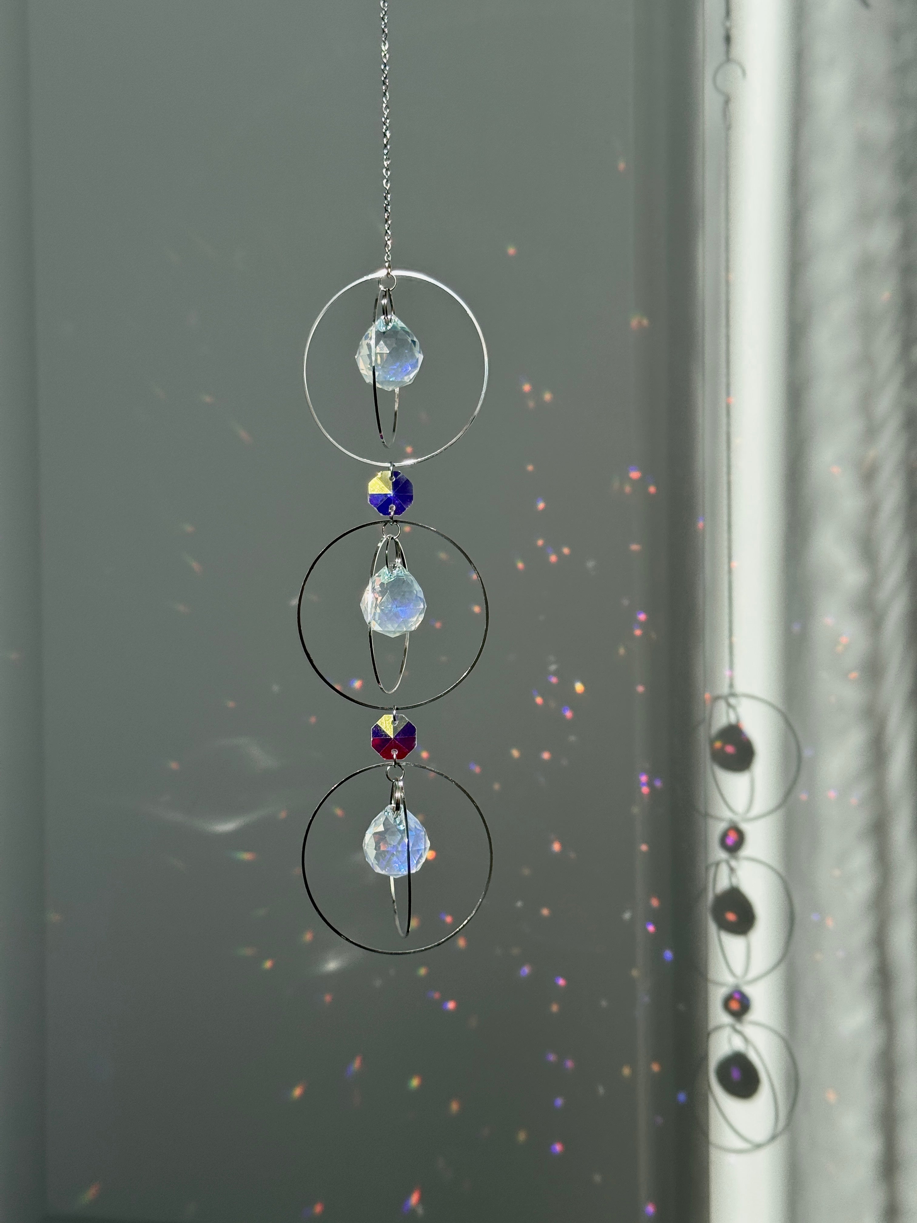 A silver-toned triple-tiered suncatcher made of stainless steel hoops. Each of the three orbits holds a clear crystal sphere with an AB coating, reflecting iridescent rainbow colors against a neutral background.