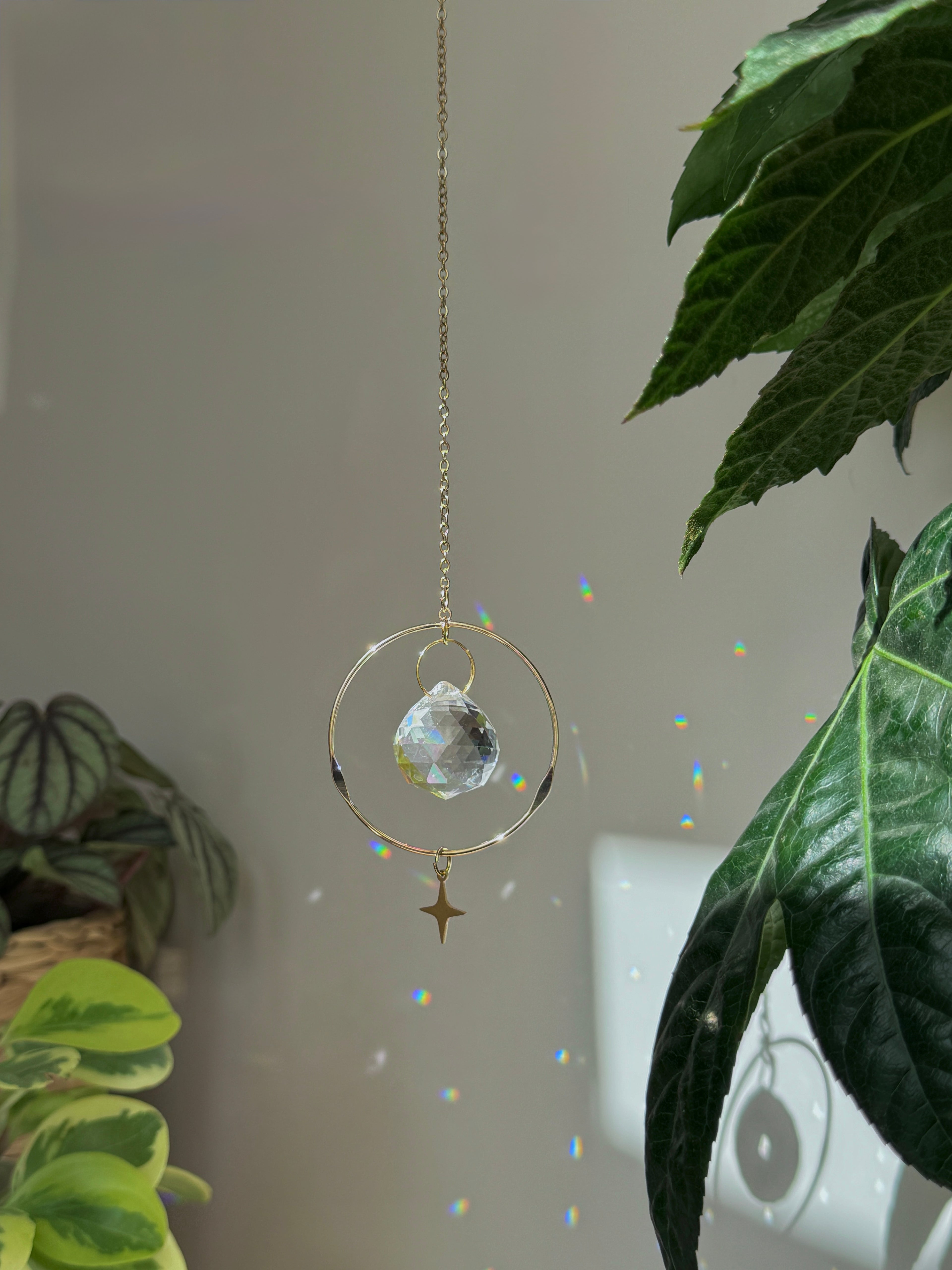 Celestial Compass suncatcher (Sonnenfänger) with a gold wavy brass ring,  crystal prism, and star charm, reflecting rainbows (Regenbogen) on a white wall.