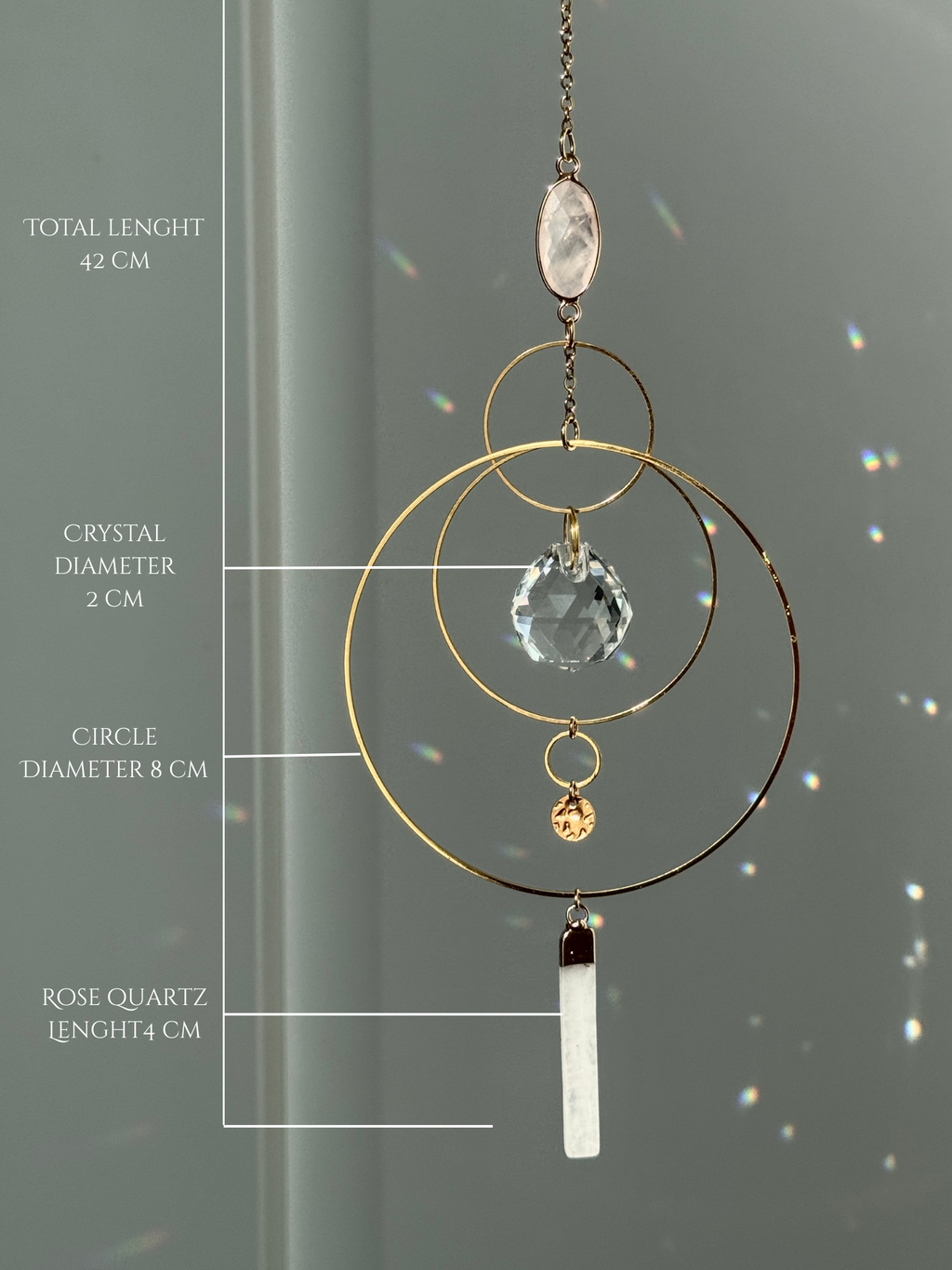 A gold geometric suncatcher featuring two pink Rose Quartz gemstones and a faceted crystal sphere. The suncatcher consists of two interlocking gold hoops, a textured coin charm, and is hanging against a soft-focus background, reflecting bright light.