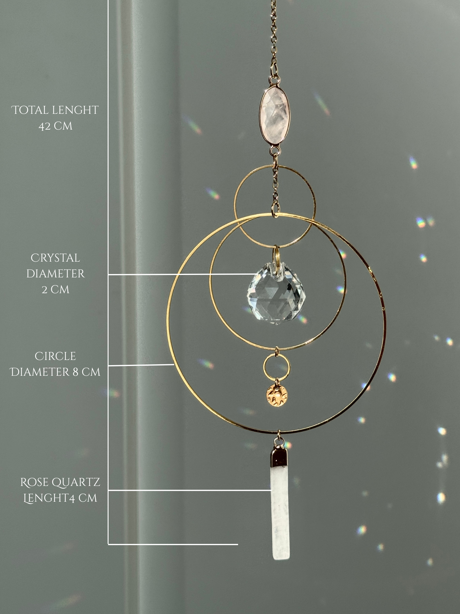 A gold geometric suncatcher featuring two pink Rose Quartz gemstones and a faceted crystal sphere. The suncatcher consists of two interlocking gold hoops, a textured coin charm, and is hanging against a soft-focus background, reflecting bright light.