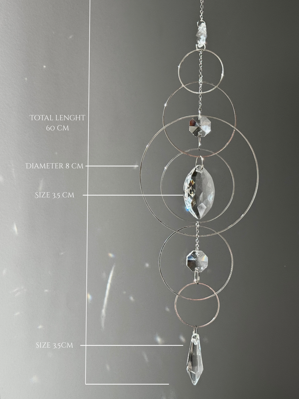 60cm silver geometric suncatcher featuring interlocking concentric hoops and clear marquise-cut crystal prisms, hanging in a window and casting rainbows on a wall.