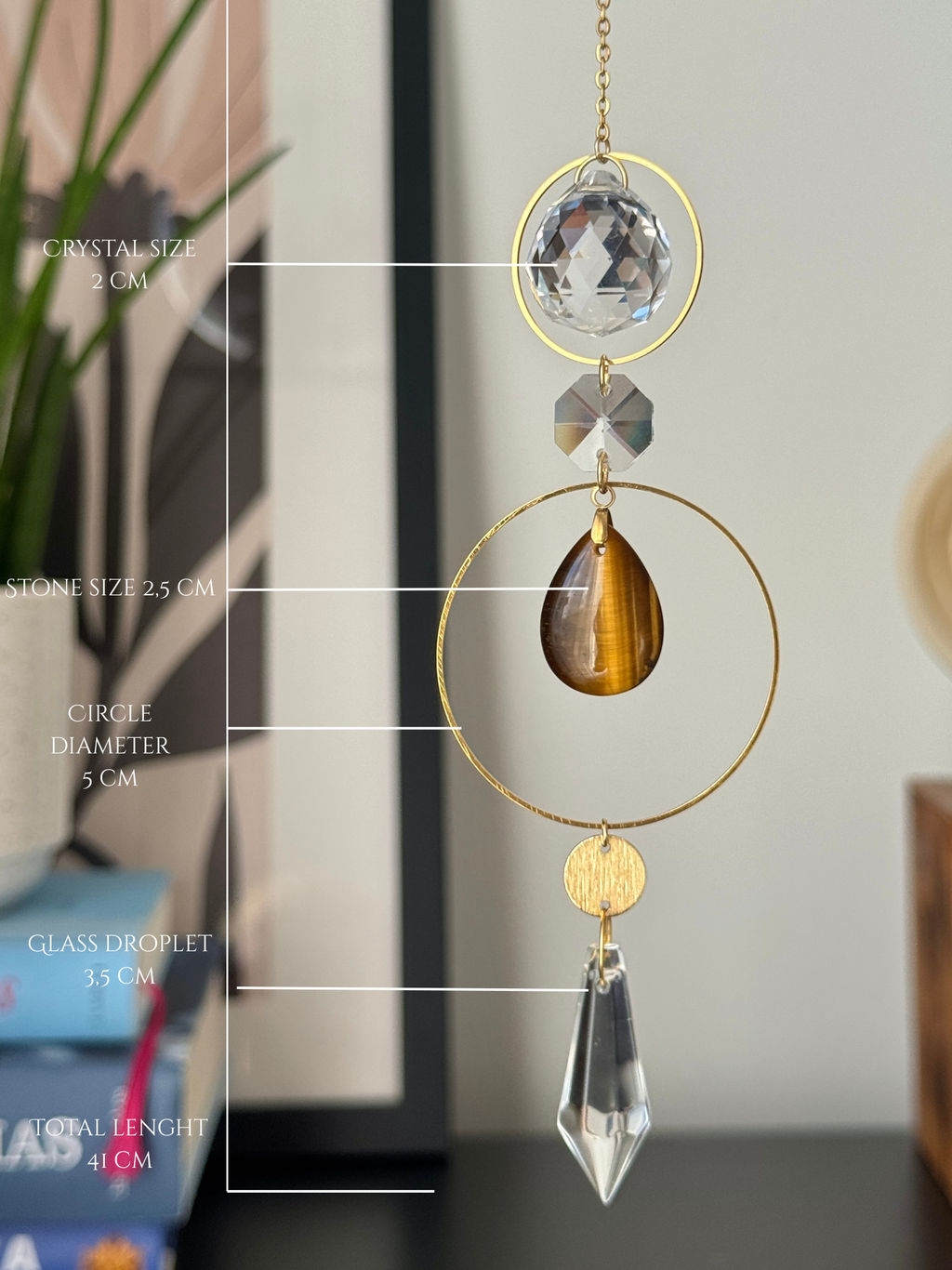 A gold suncatcher ornament with a clear crystal, a brown tiger's eye teardrop, and a clear glass droplet, shown with white measurement lines and text indicating dimensions.