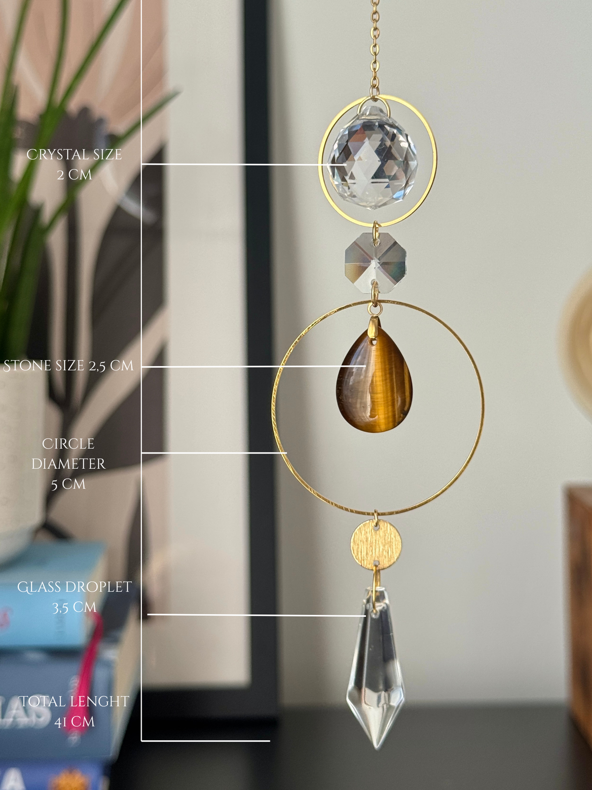 A gold suncatcher ornament with a clear crystal, a brown tiger's eye teardrop, and a clear glass droplet, shown with white measurement lines and text indicating dimensions.