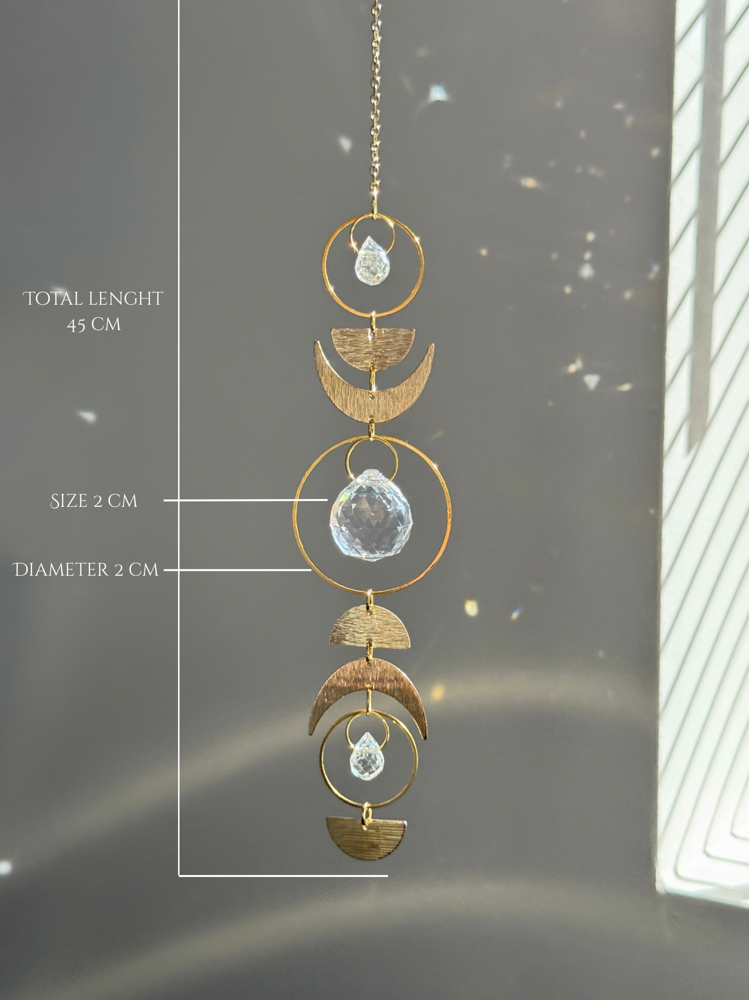 Handcrafted gold moon phase suncatcher featuring a vertical alignment of textured brass crescent moons and hoops with three clear crystal prisms casting rainbows on a white wall.